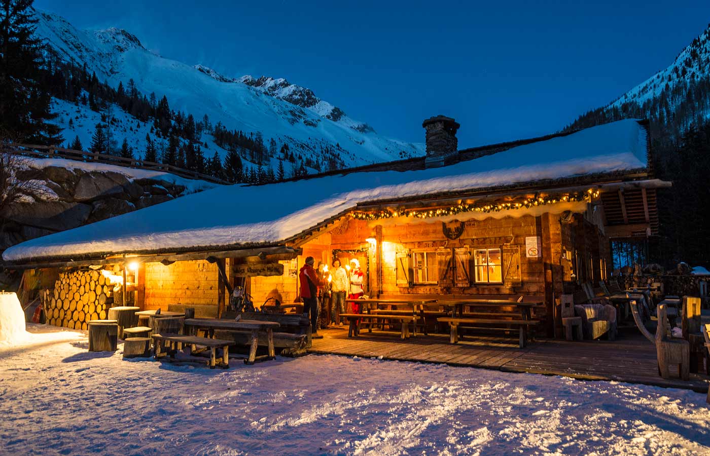 View of a wooden alpine hut illuminated at night with snow on the roof and three people speaking out of the door