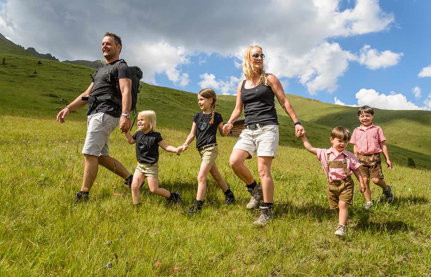 Famiglia con padre e madre che tengono per mano quattro bambini passeggia sui pascoli verdi di montagna