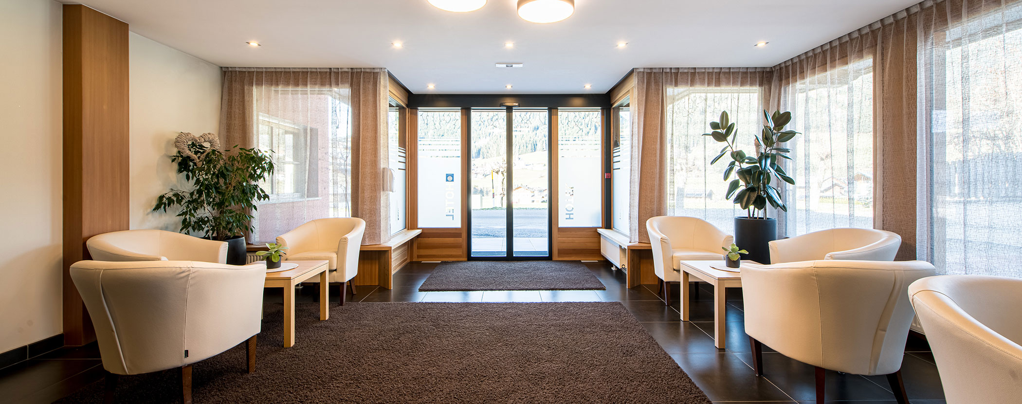 Helles, modernes Wartezimmer mit weißen Stühlen, Pflanzen, großen Fenstern und gläsernen Eingangstüren. - Hotel Waldheim
