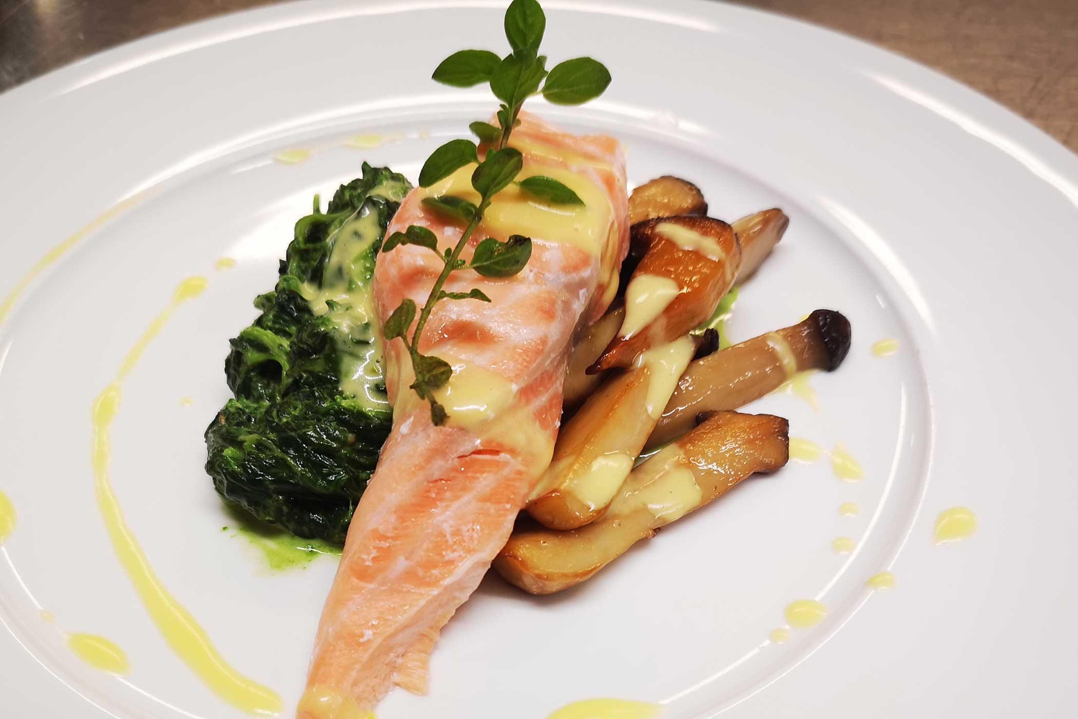 Lachsfilet mit Sahnesauce, sautierten Pilzen und sautiertem Spinat, garniert mit frischen Kräutern auf einem weißen Teller. - Hotel Waldheim