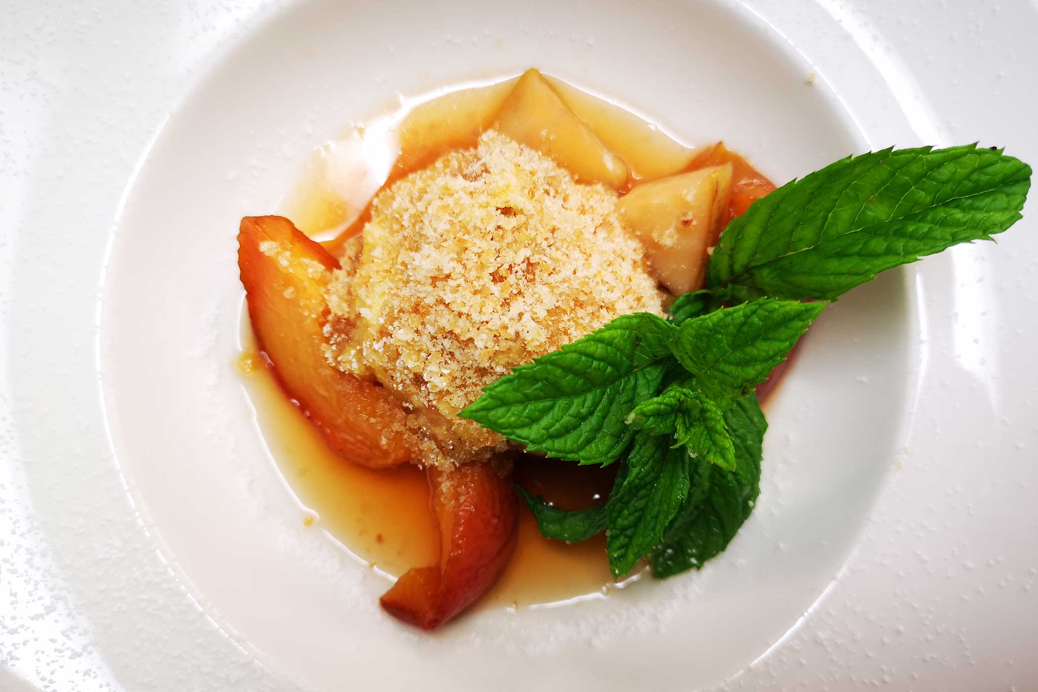 Ein Dessert mit Pfirsichscheiben, Krümelbelag, Sauce und frischer Minze auf einem weißen Teller. - Hotel Waldheim