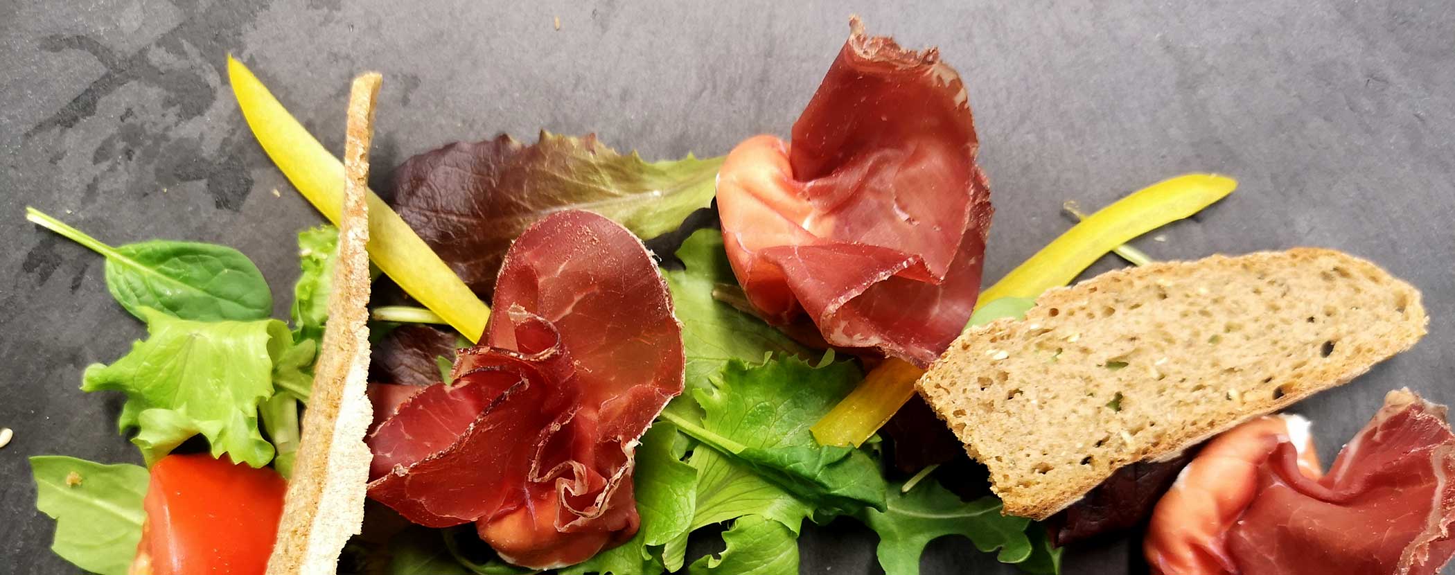 Gemischtes Grünzeug mit gepökeltem Fleisch, gelben Paprikastreifen, Tomaten und Brotscheiben auf einer dunklen Unterlage. - Hotel Waldheim