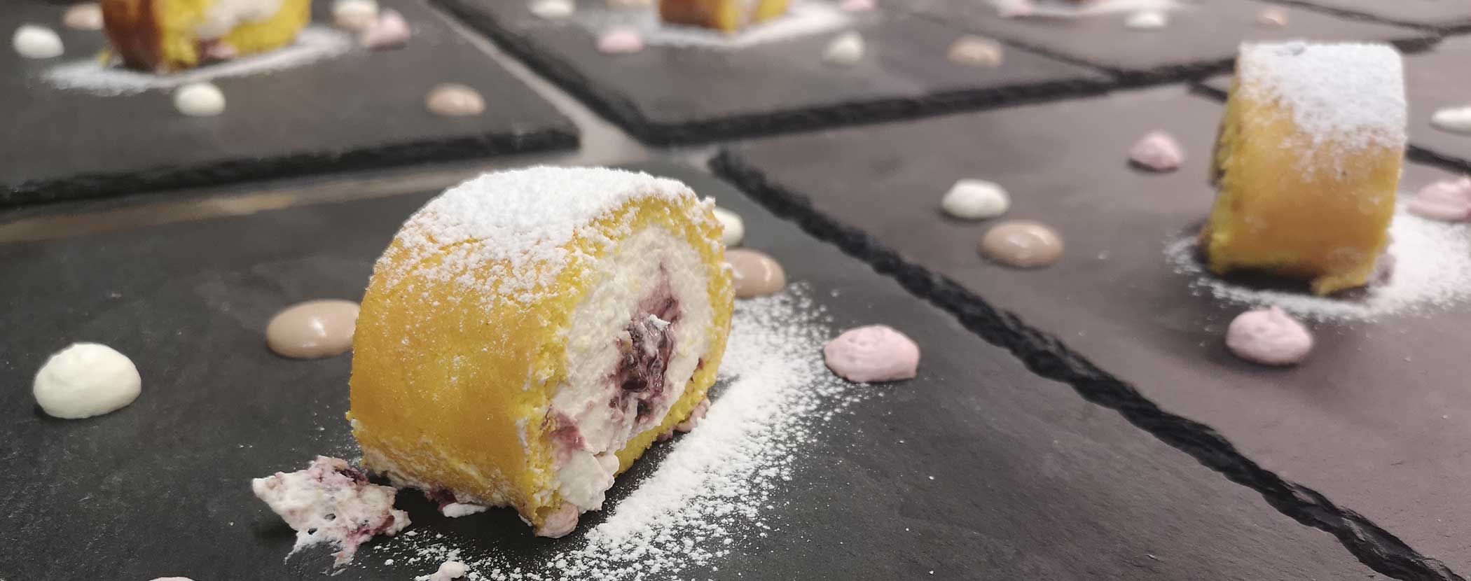 Ein in Scheiben geschnittenes Dessertbrötchen mit Puderzucker, umgeben von kleinen Sahnetupfern auf einem Schiefertablett. - Hotel Waldheim