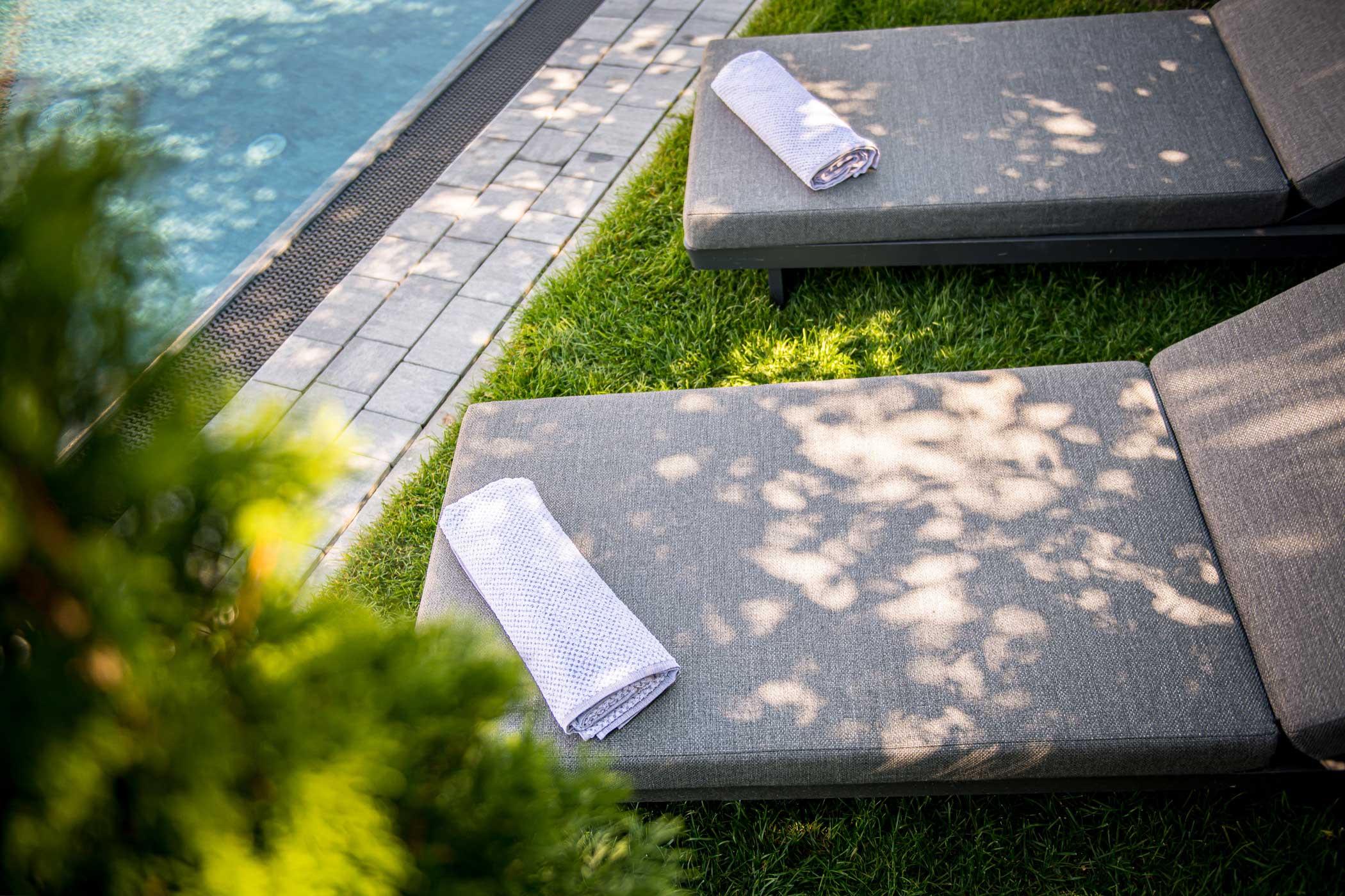 Zwei graue Liegestühle mit aufgerollten weißen Handtüchern neben einem Pool, beschattet von Baumzweigen. - Hotel Waldheim