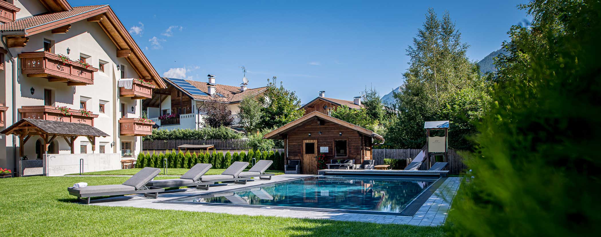 Ein luxuriöser Außenpool mit Liegestühlen, umgeben von Gras und Gebäuden im alpinen Stil unter einem klaren blauen Himmel. - Hotel Waldheim