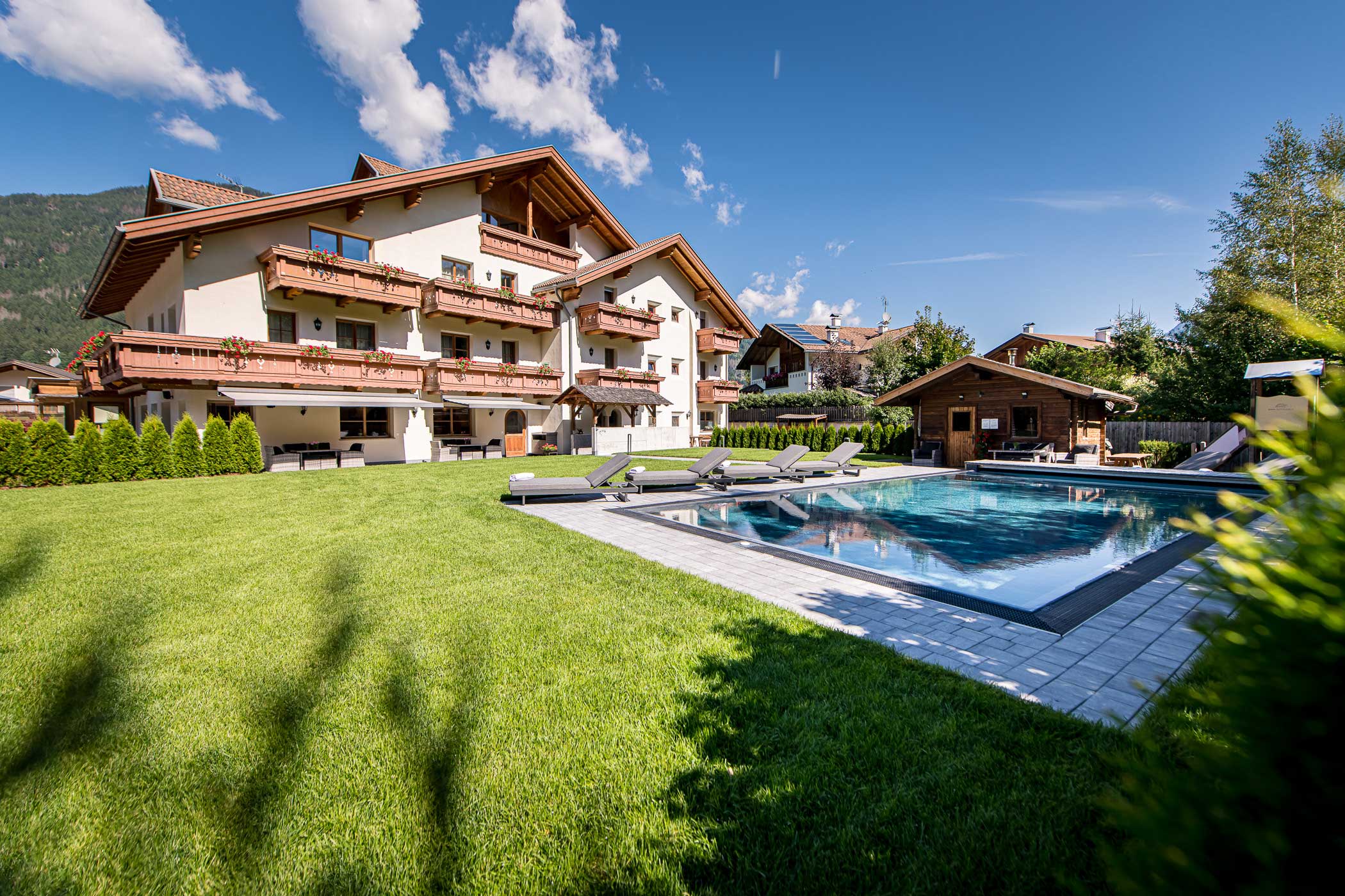 Großes Haus im alpinen Stil mit Balkonen, Swimmingpool, Liegewiese, Liegestühlen und Bergen im Hintergrund. - Hotel Waldheim