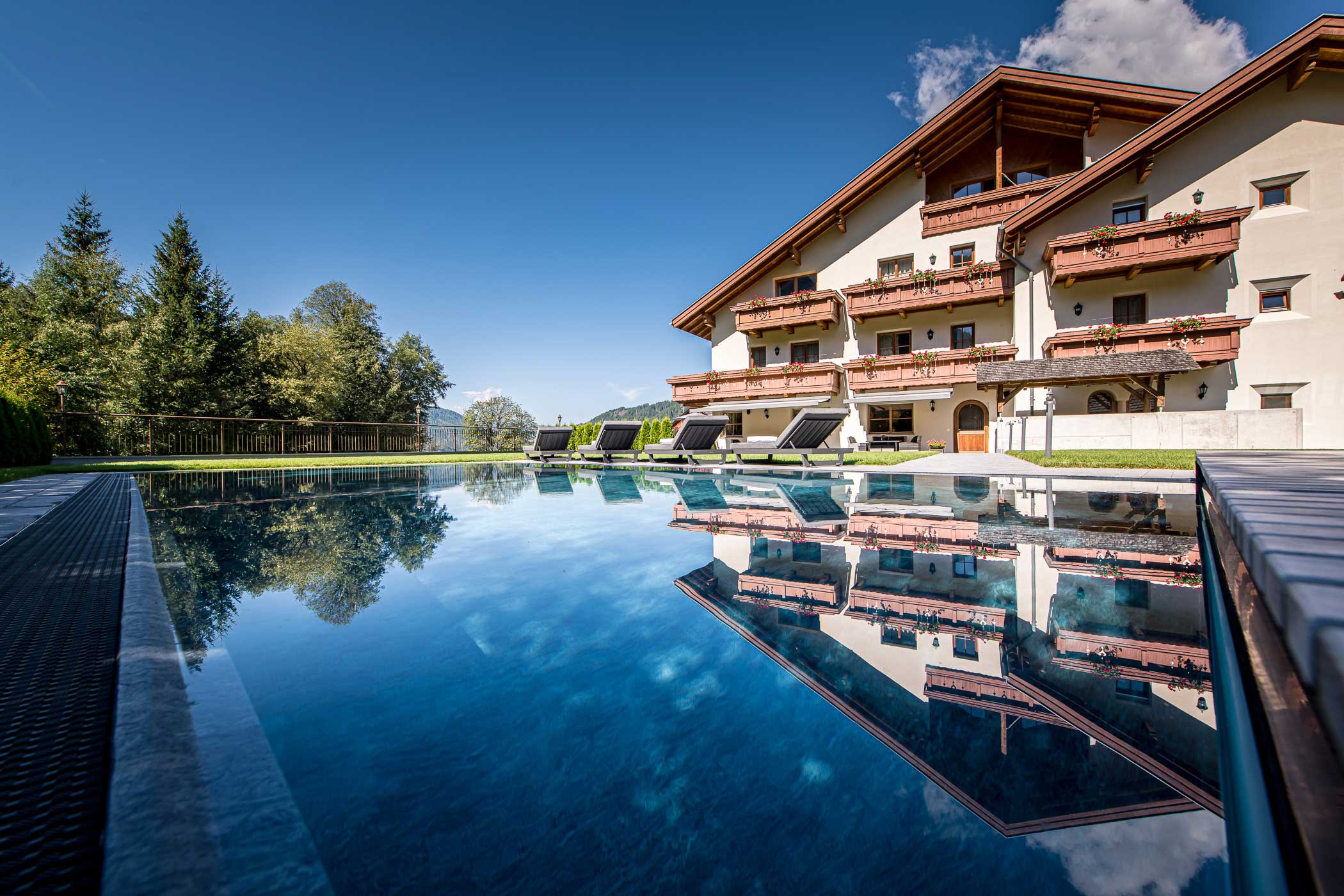 Ein großes Hotel im Chalet-Stil, das sich in einem klaren Außenpool unter einem strahlend blauen Himmel spiegelt. - Hotel Waldheim