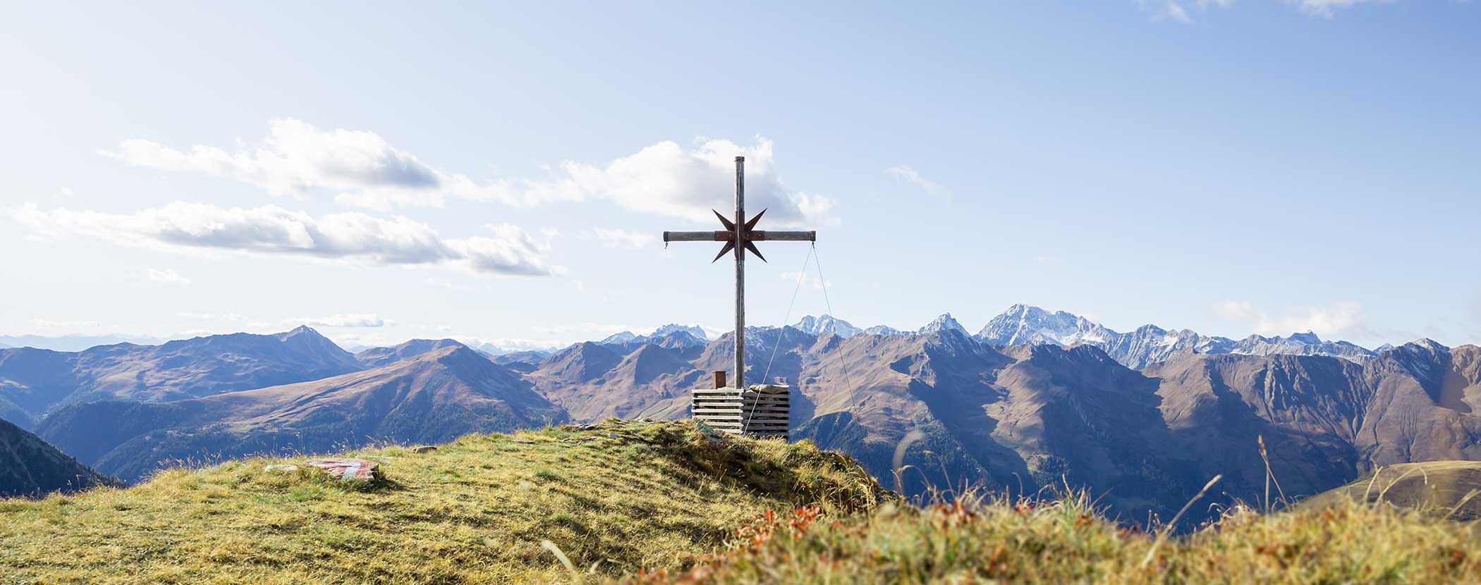 Ein Holzkreuz steht auf einem grasbewachsenen Hügel mit fernen Bergen und einem blauen Himmel im Hintergrund. - Hotel Waldheim