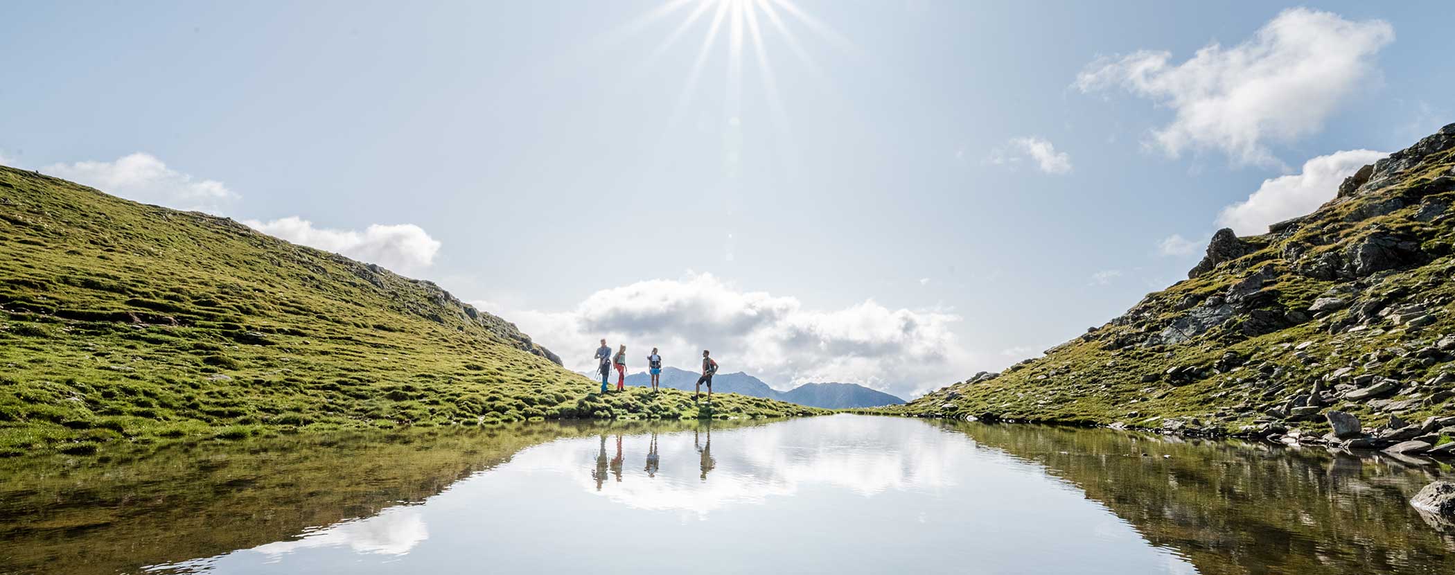 Vier Wanderer spazieren an einem spiegelnden Bergsee unter einem sonnigen, blauen Himmel mit vereinzelten Wolken. - Hotel Waldheim