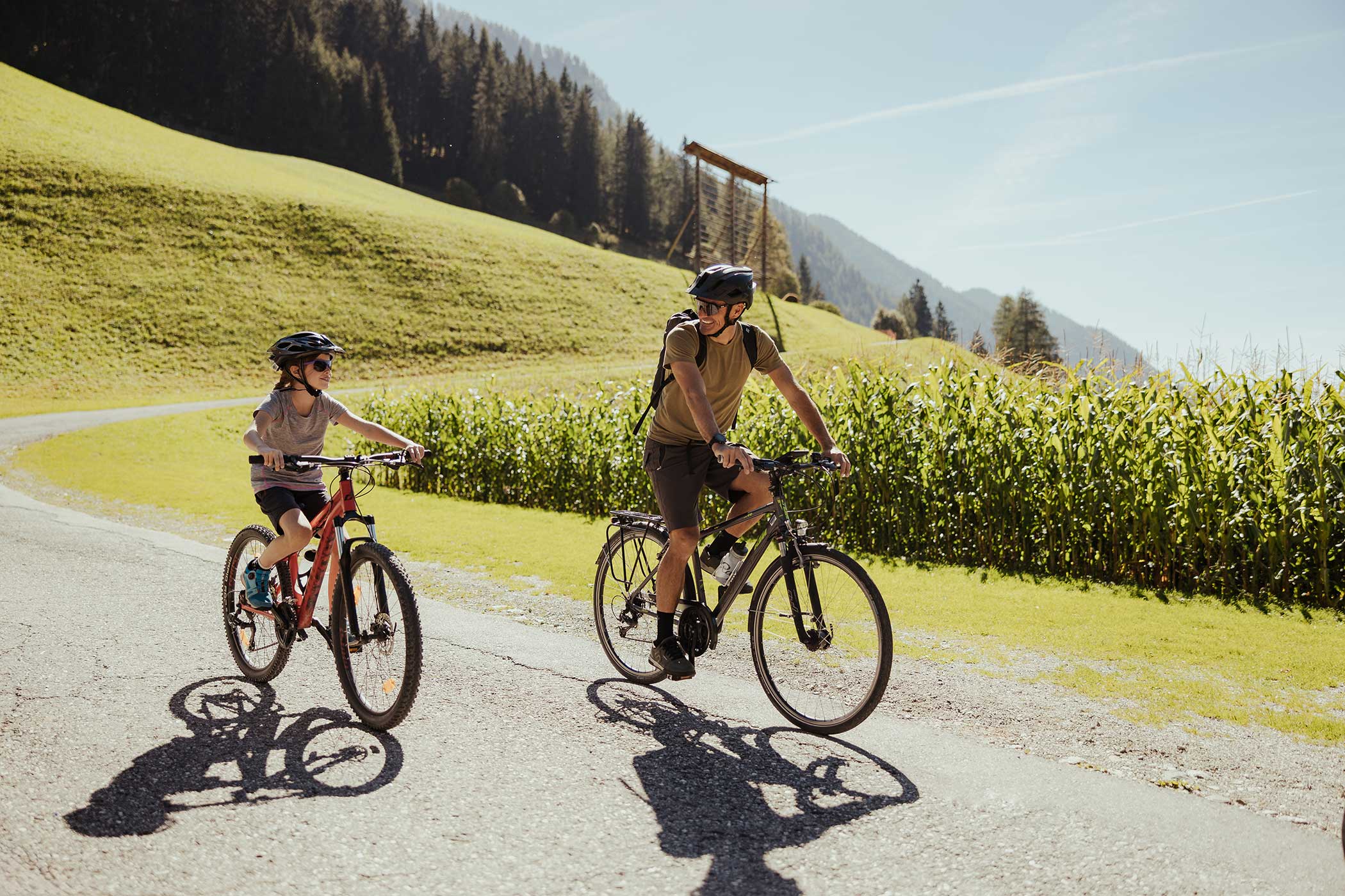 Ein Erwachsener und ein Kind fahren mit dem Fahrrad auf einer sonnigen Landstraße mit grünen Hügeln und Bäumen im Hintergrund. - Hotel Waldheim