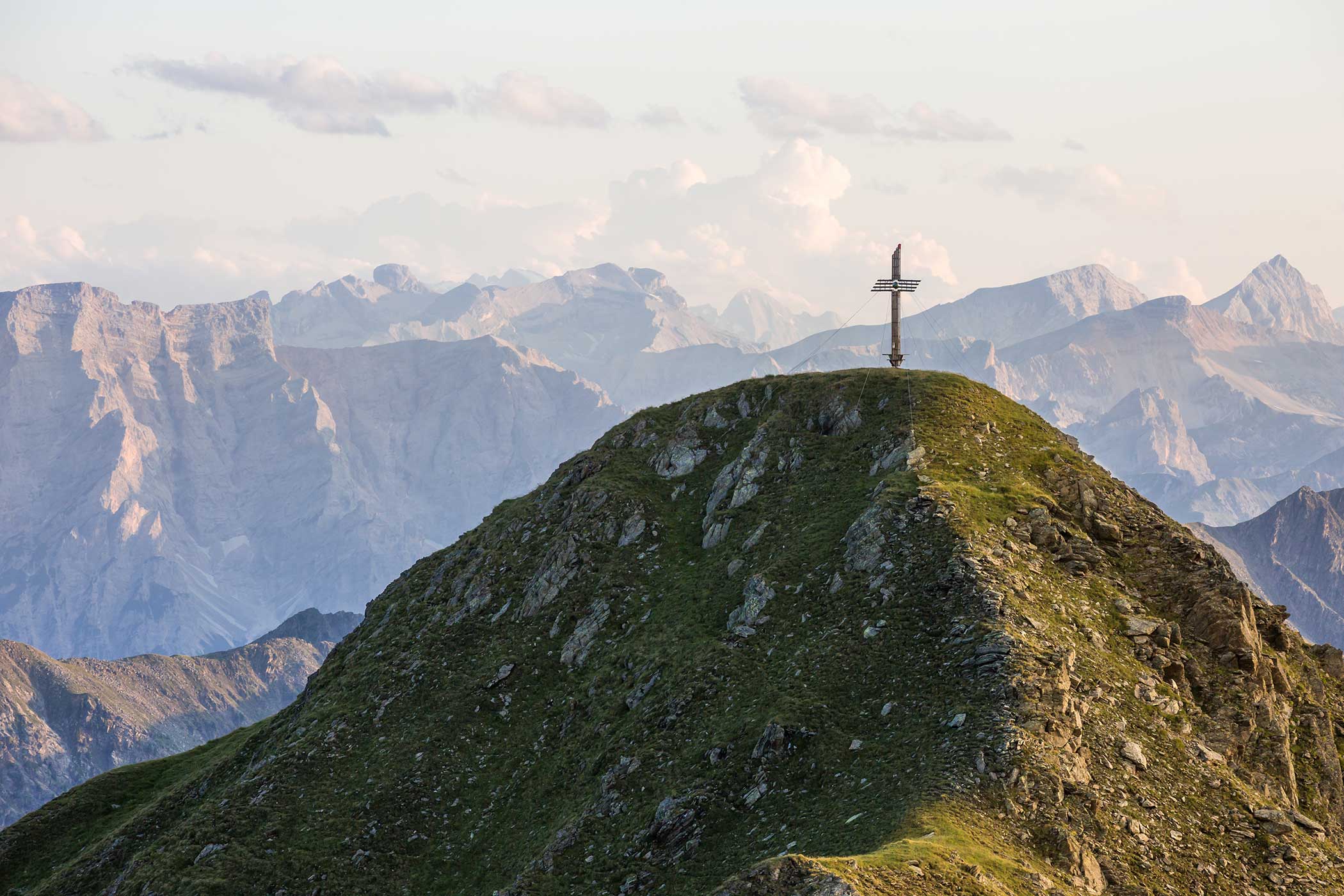 Ein großes Kreuz steht auf einem grasbewachsenen Berggipfel mit schroffen Bergen im Hintergrund. - Hotel Waldheim