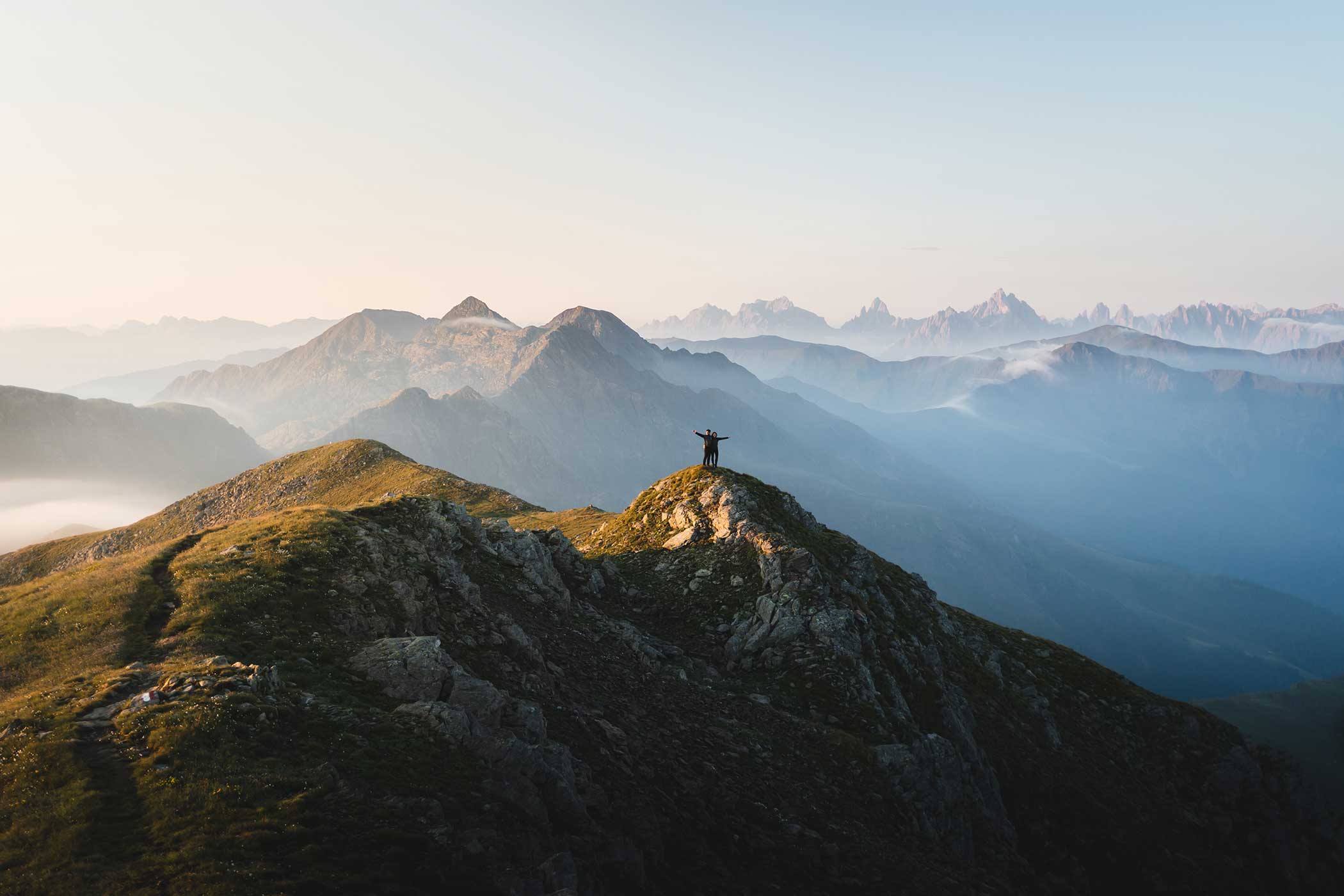 Person, die mit erhobenen Armen auf einem Berggipfel steht, umgeben von nebligen Bergketten bei Sonnenaufgang. - Hotel Waldheim