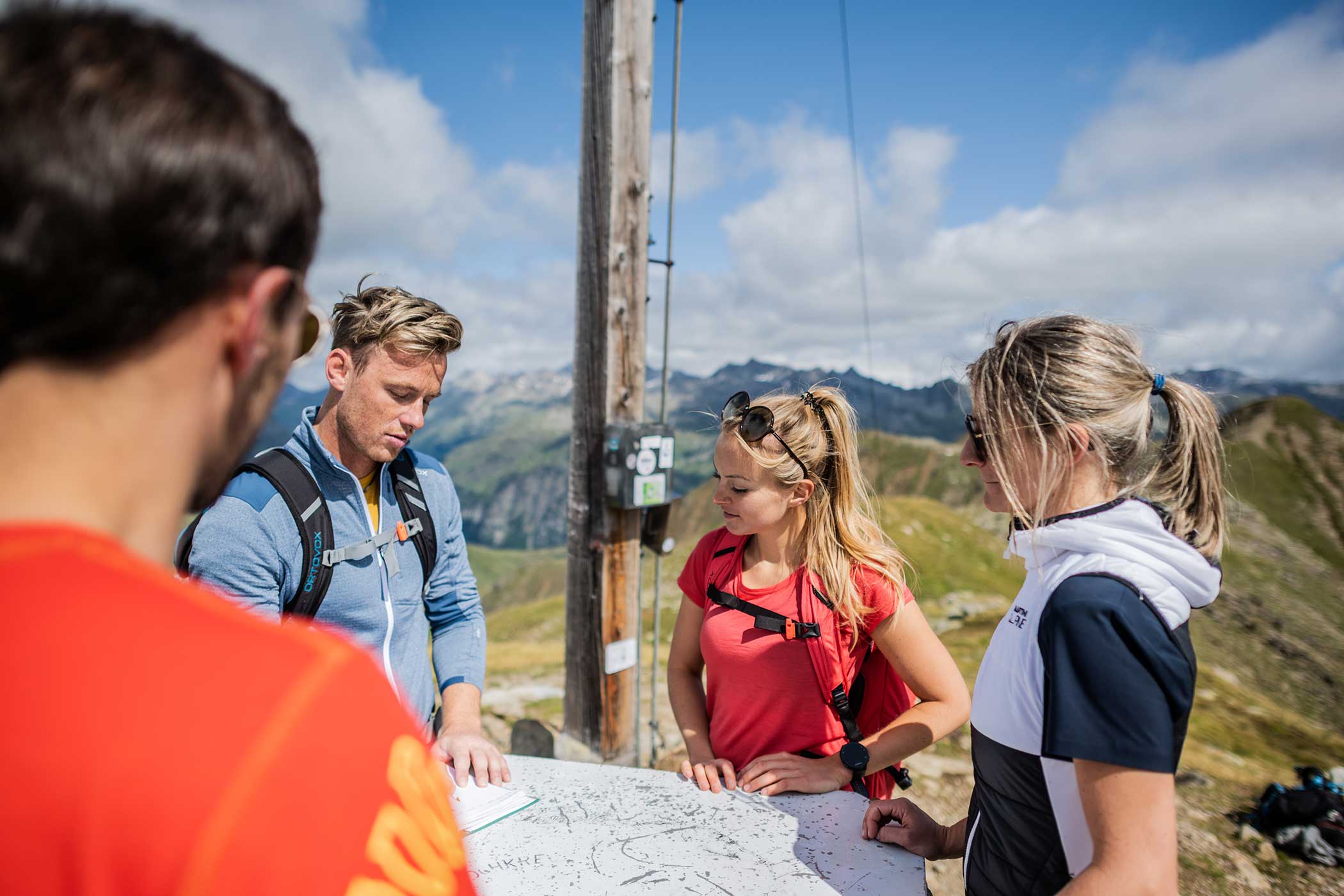 Vier Wanderer studieren an einem sonnigen Tag an einem Aussichtspunkt auf einem Berggipfel eine Karte, mit Bergen im Hintergrund. - Hotel Waldheim