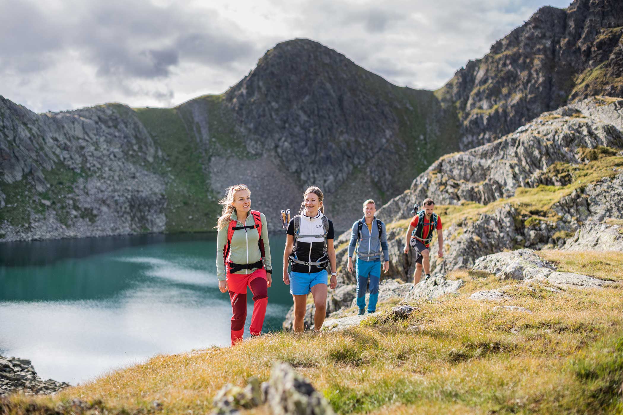 Vier Personen wandern auf einem grasbewachsenen Bergpfad an einem blauen See, umgeben von felsigen Gipfeln unter einem bewölkten Himmel. - Hotel Waldheim