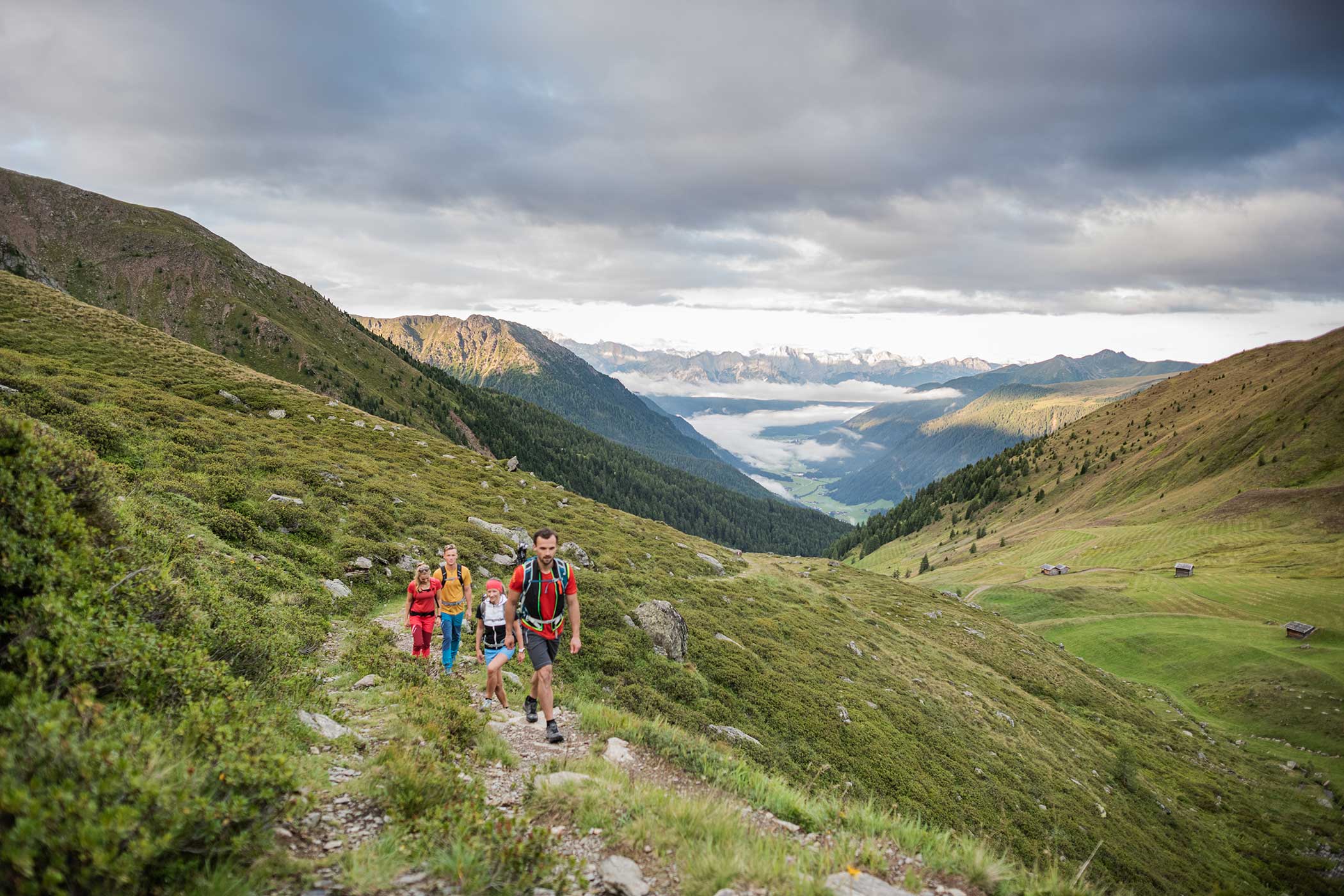 Eine Familie wandert auf einem grasbewachsenen Bergpfad mit einem malerischen Tal und fernen Bergen unter einem bewölkten Himmel. - Hotel Waldheim