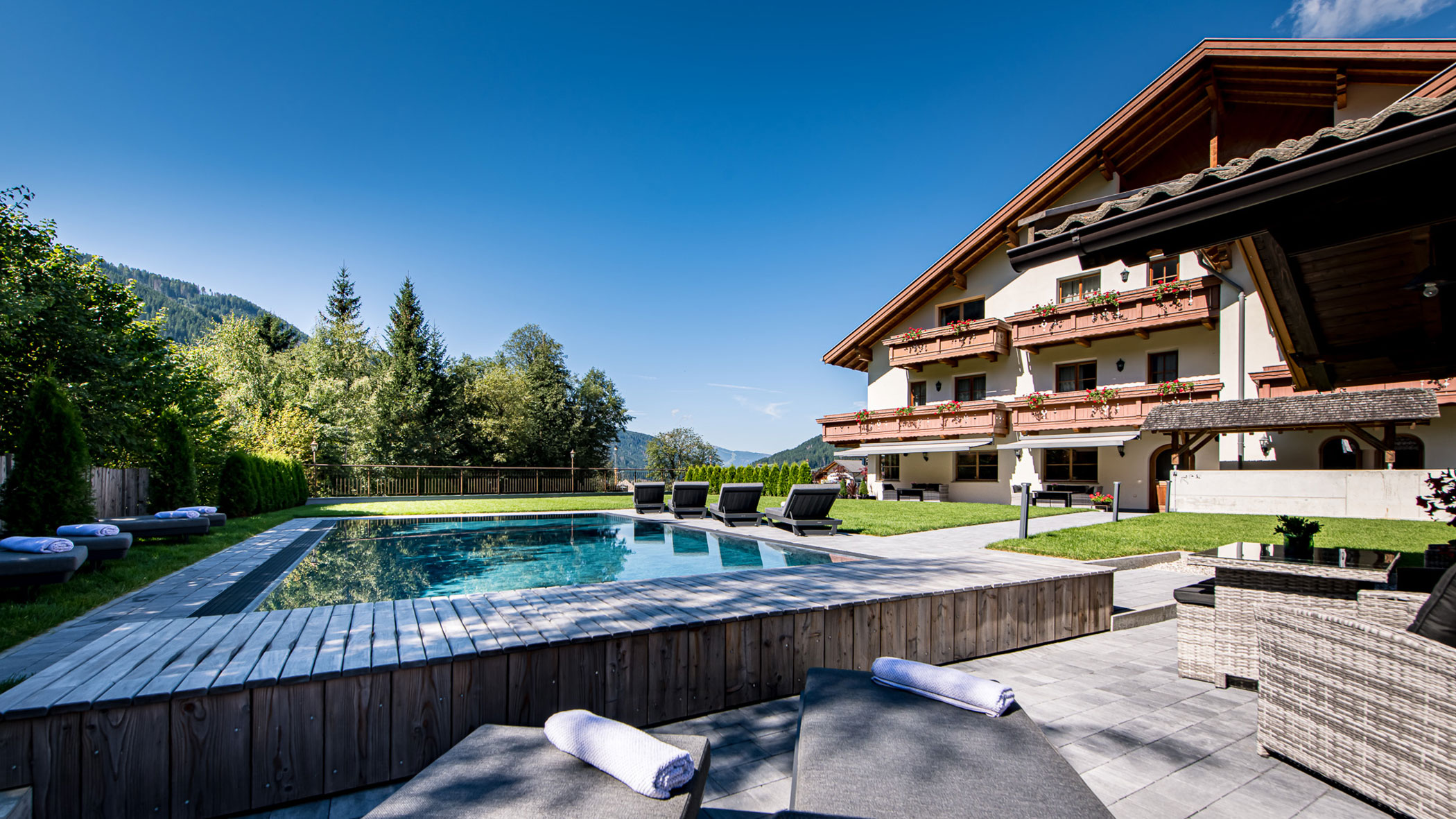 Außenpool mit Liegestühlen neben einem Hotel im Chalet-Stil, umgeben von Bäumen und Bergen unter blauem Himmel. - Hotel Waldheim