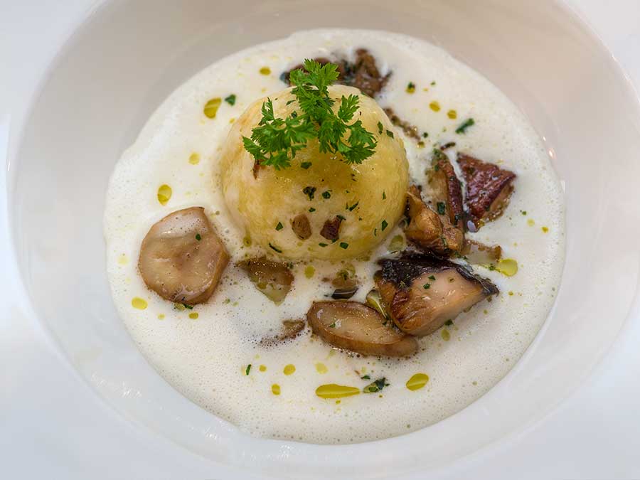 Ein Semmelknödel mit Petersilie, Pilzen und Schaumsauce, garniert mit Kräutern und Öltropfen in einer weißen Schale. - Hotel Waldheim