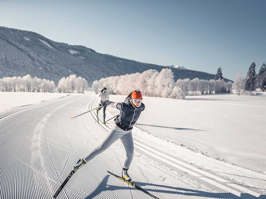 Zwei Personen beim Skilanglauf auf einer präparierten, verschneiten Loipe mit Bergen und Bäumen im Hintergrund. - Hotel Waldheim