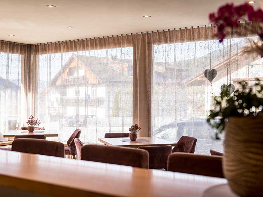 Gemütliches Café mit Tischen, Topfpflanzen und großen Fenstern, durch die man die Häuser und die Berge sehen kann. - Hotel Waldheim