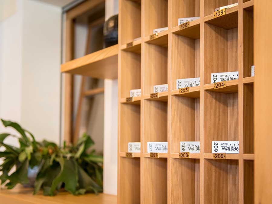 Holzfächer für Hotelschlüssel mit beschrifteten Schlüsselkarten und eine Grünpflanze auf einem Regal in der Nähe. - Hotel Waldheim