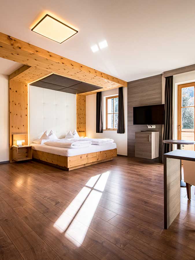 Modernes Hotelzimmer mit Holzboden, großem Bett, Fernseher und Sonnenlicht, das durch die Fenster fällt. - Hotel Waldheim