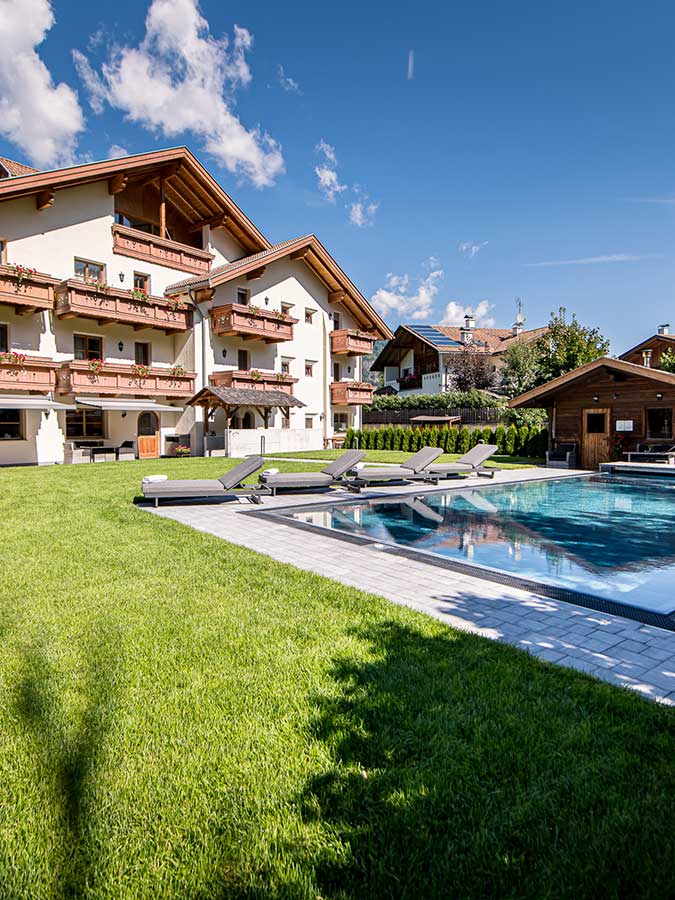 Sonniger Blick auf ein modernes Haus im Chalet-Stil mit Balkonen und einem Pool, umgeben von grünem Rasen und Liegestühlen. - Hotel Waldheim