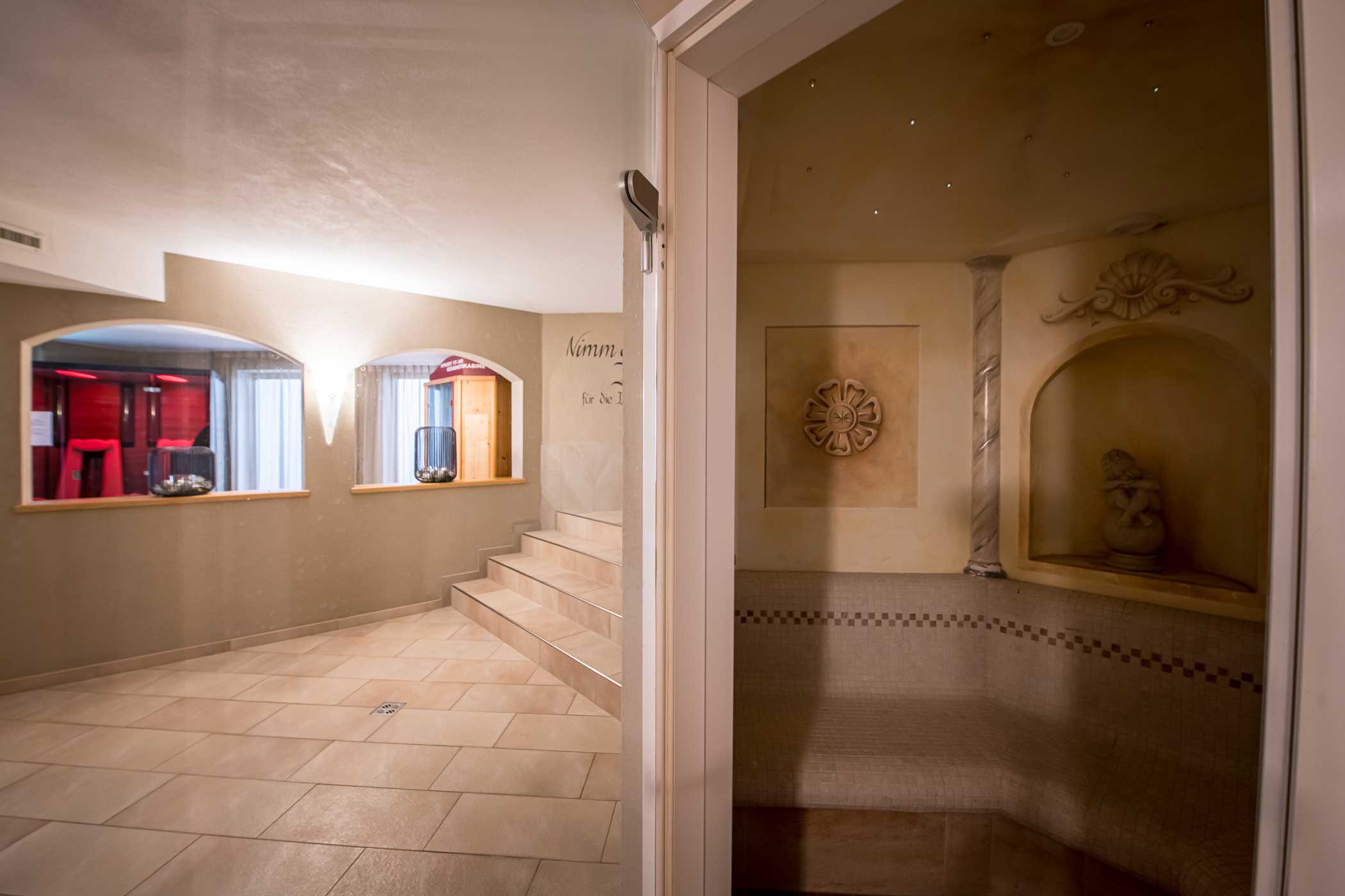 Spa-Bereich mit gefliesten Böden, Stufen, links ein Saunaraum und rechts eine dekorative Nische mit einer Statue. - Hotel Waldheim