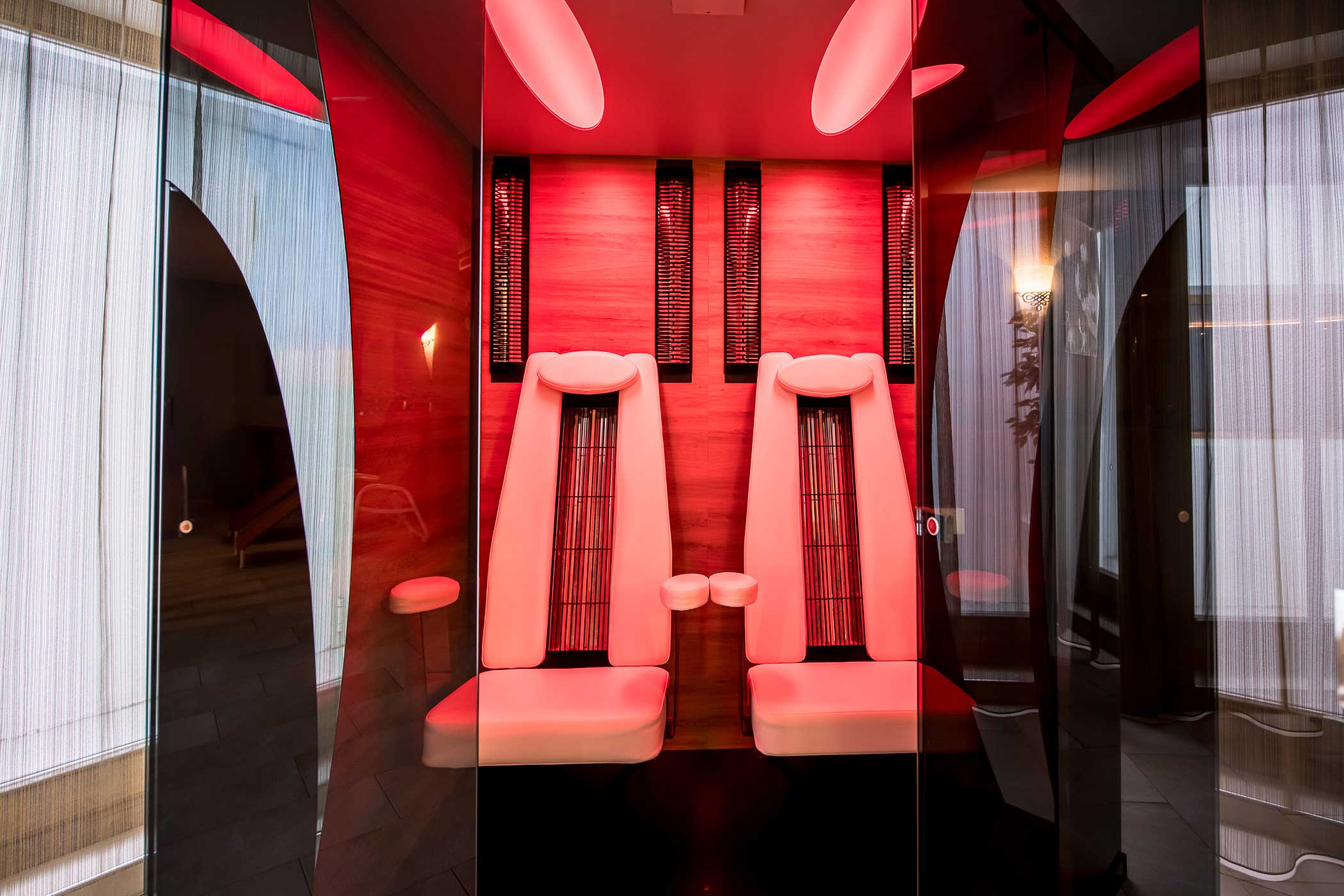 Zwei moderne Loungesessel in einem Wellnessbereich mit roter Umgebungsbeleuchtung und vertikalen Heizkörpern hinter den Sesseln. - Hotel Waldheim