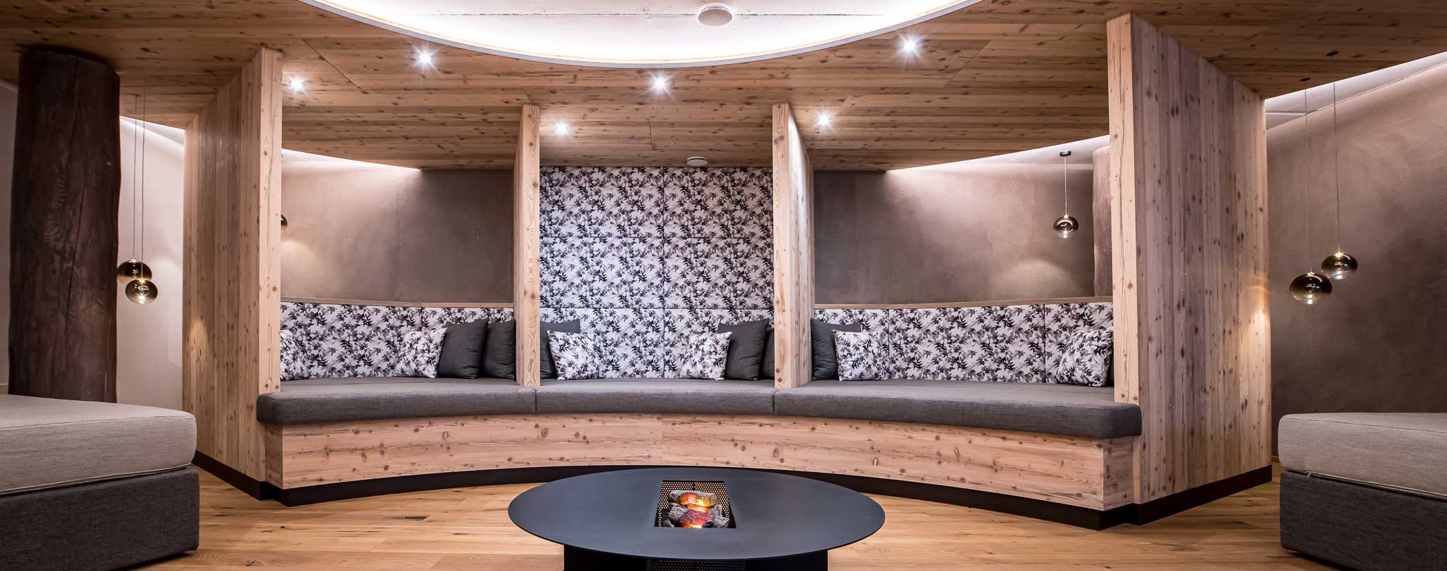 Moderne Lounge mit Holzkabinen, gemusterten Kissen und einem runden Tisch mit eingebauter Heizung in der Mitte. - Hotel Waldheim