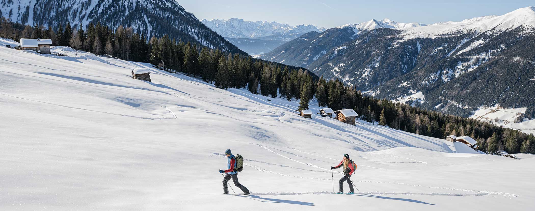 Zwei Personen fahren bergauf durch verschneite Berge mit verstreuten Hütten und Wäldern unter einem klaren blauen Himmel. - Hotel Waldheim