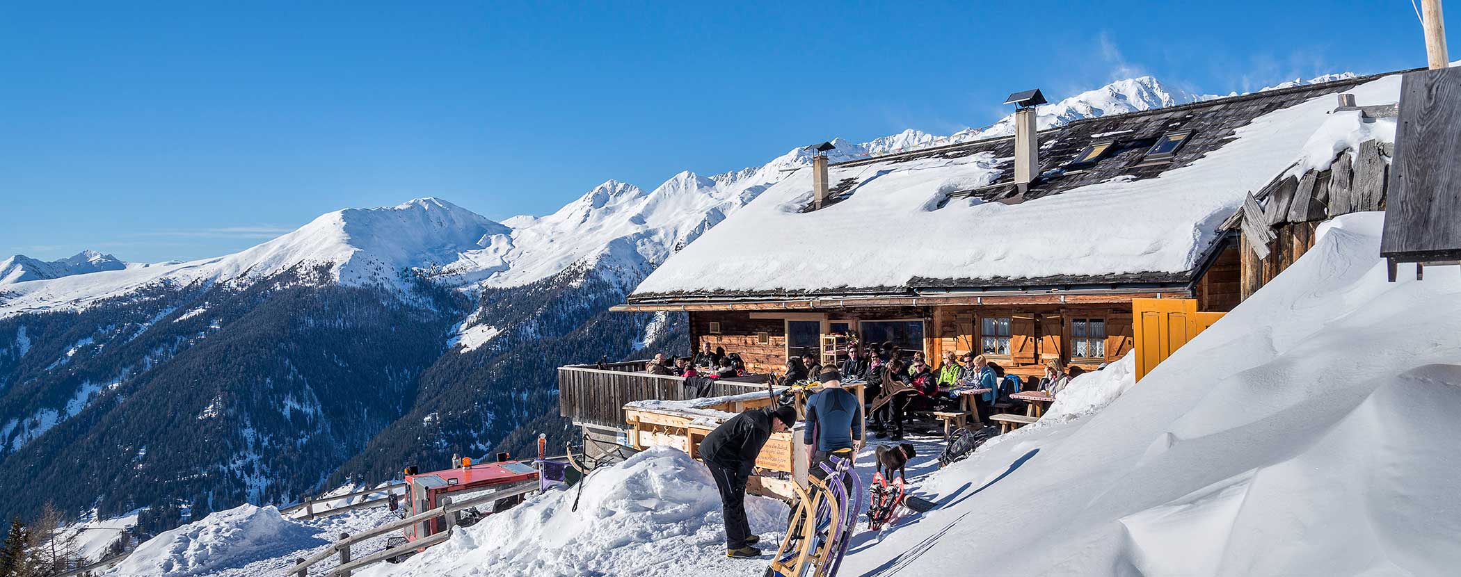 Menschen, die einen sonnigen Tag vor einer verschneiten Berghütte mit Skipisten und schneebedeckten Gipfeln im Hintergrund genießen. - Hotel Waldheim