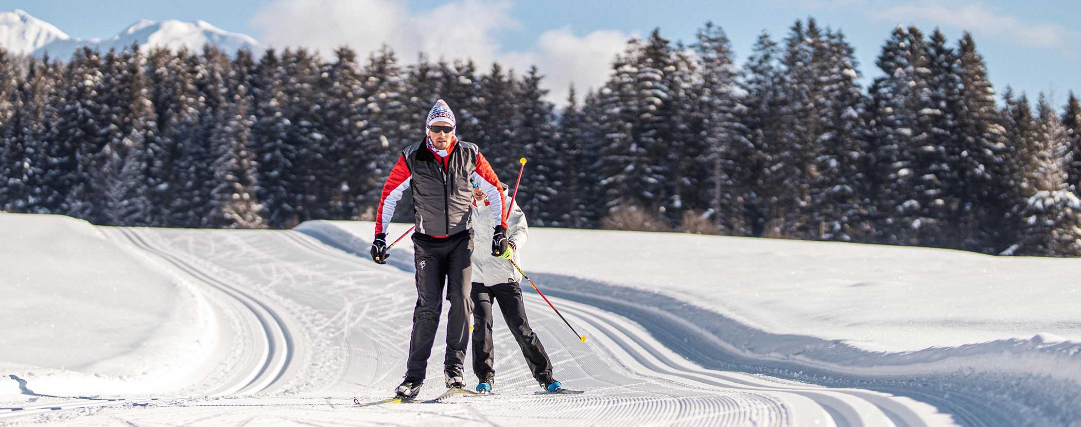 Zwei Personen beim Skilanglauf auf einer verschneiten Loipe mit Bäumen und Bergen im Hintergrund. - Hotel Waldheim