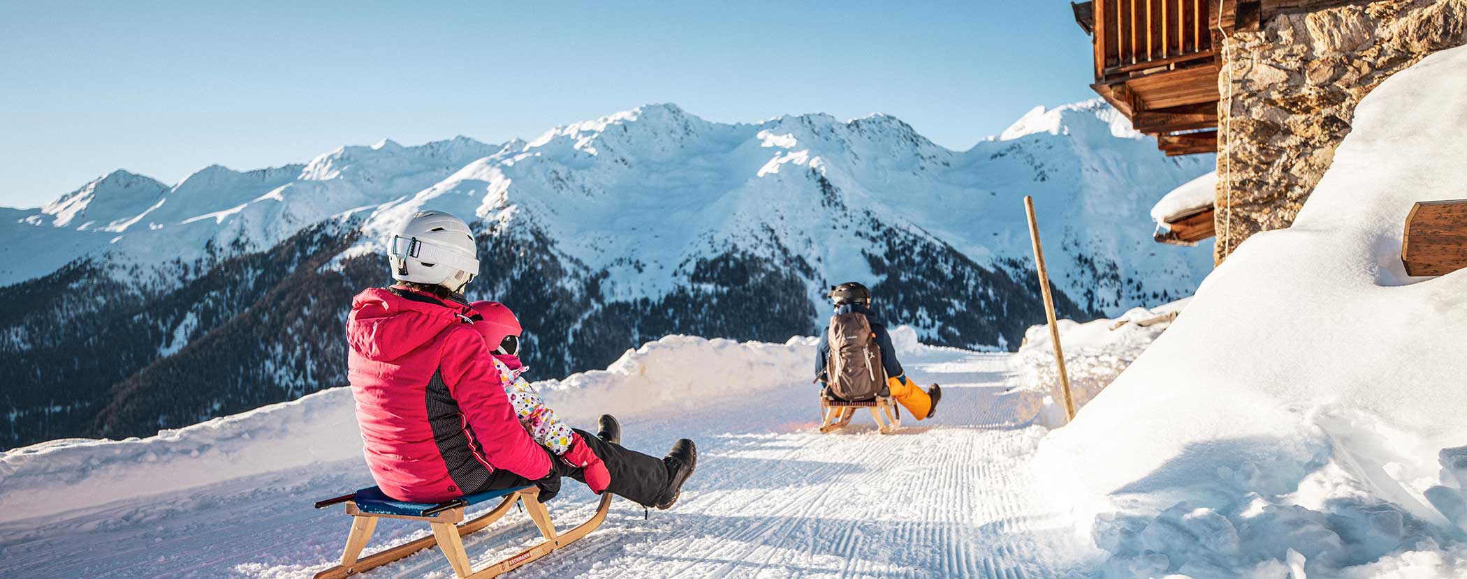 Zwei Menschen in Winterkleidung rodeln auf einem verschneiten Weg mit Bergen im Hintergrund. - Hotel Waldheim