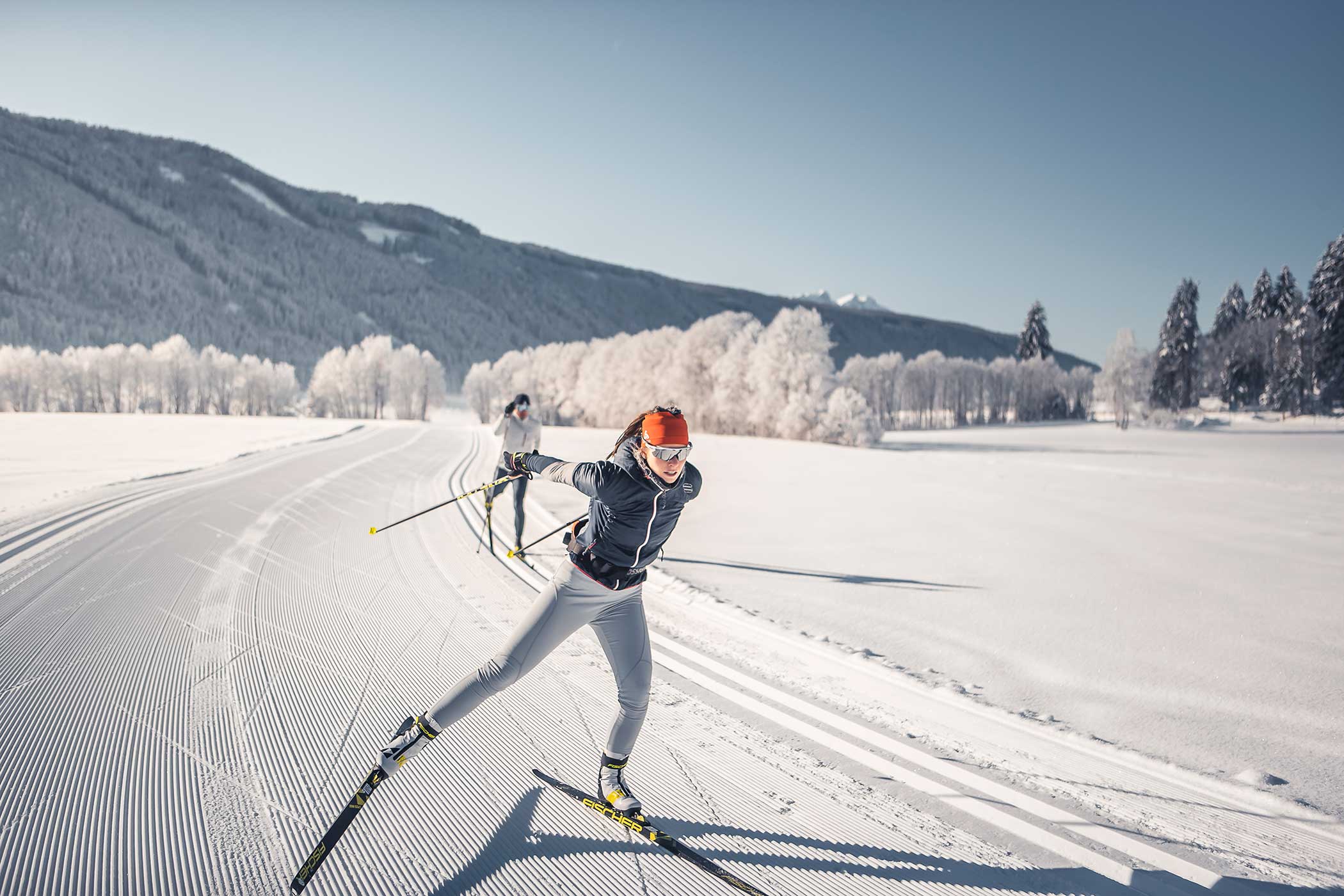 Zwei Personen beim Skilanglauf auf einer präparierten, verschneiten Loipe mit Bäumen und Bergen im Hintergrund. - Hotel Waldheim