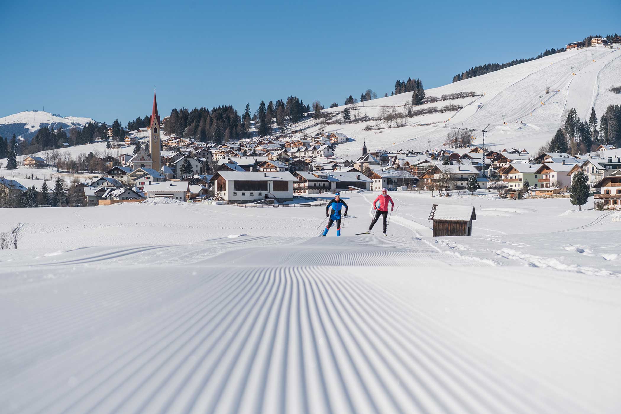 Zwei Personen beim Skilanglauf auf einer präparierten, verschneiten Loipe in der Nähe eines Dorfes mit Hügeln und Häusern im Hintergrund. - Hotel Waldheim