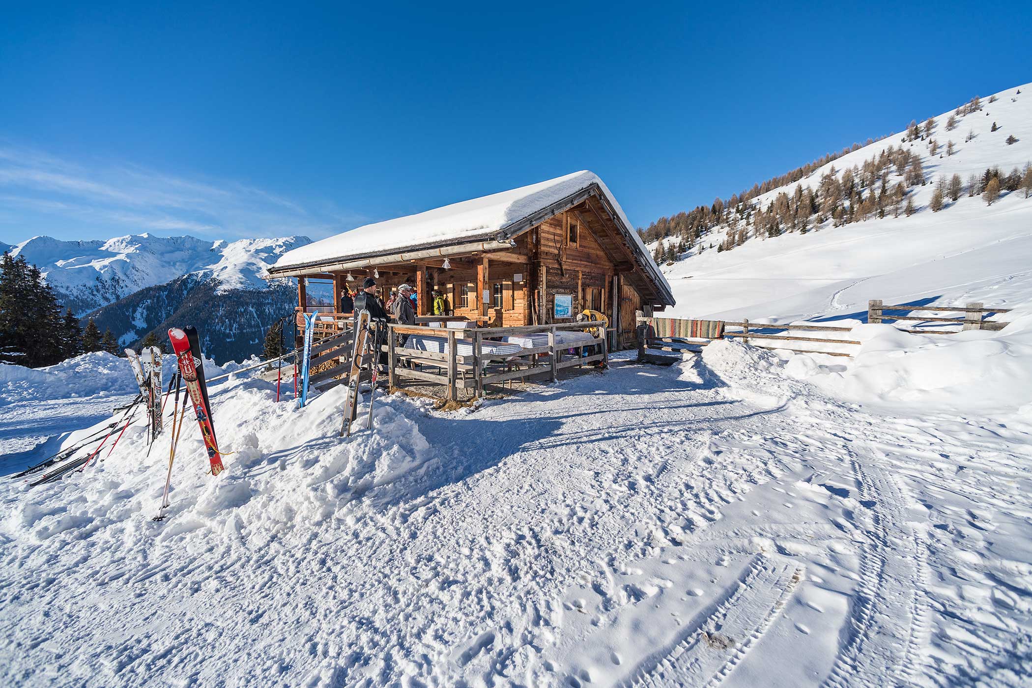 Eine hölzerne Hütte in einer verschneiten Berglandschaft mit Skiern und Stöcken unter einem klaren blauen Himmel. - Hotel Waldheim