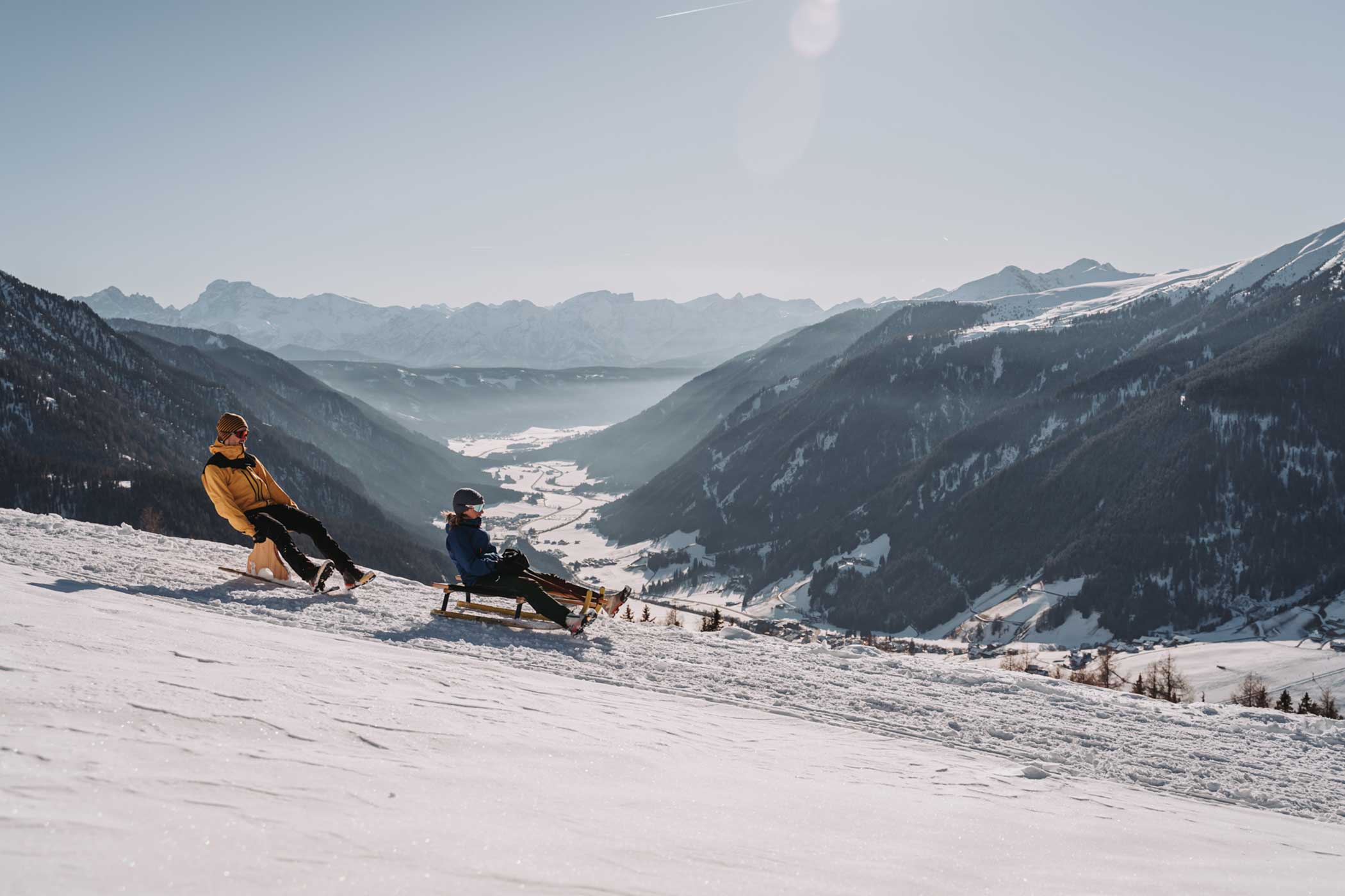 Zwei Personen fahren mit dem Schlitten einen verschneiten Berghang hinunter, mit malerischen Tälern und Bergen im Hintergrund. - Hotel Waldheim