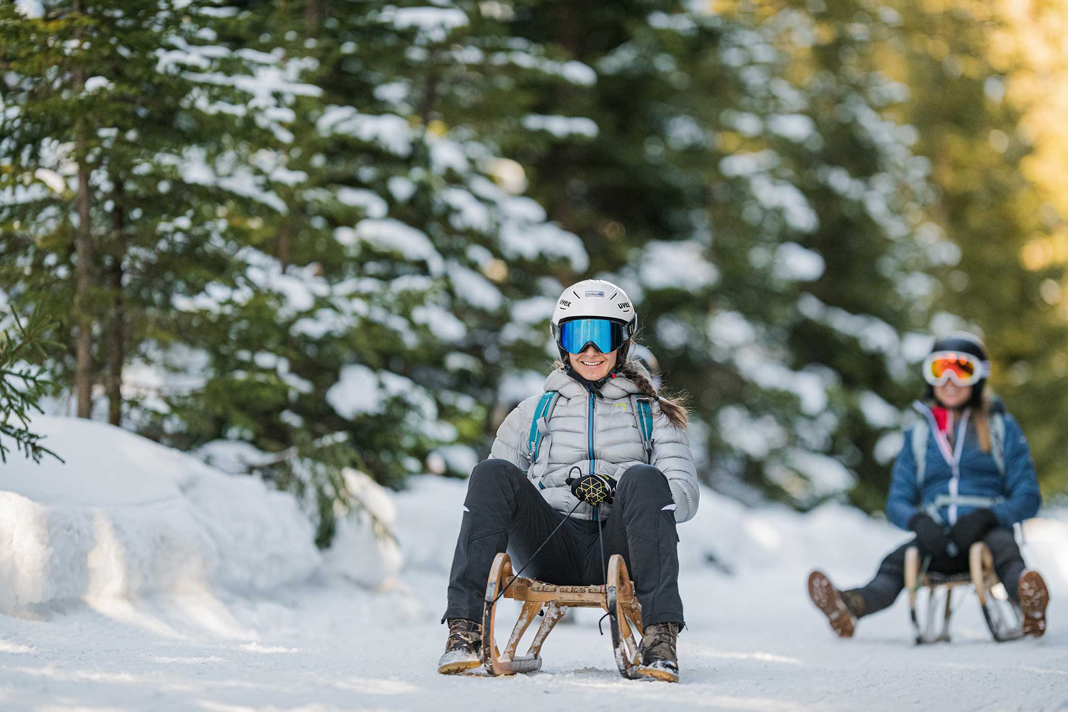 Zwei Personen fahren mit Helm, Brille und Winterkleidung auf einem verschneiten Weg durch den Wald Schlitten. - Hotel Waldheim