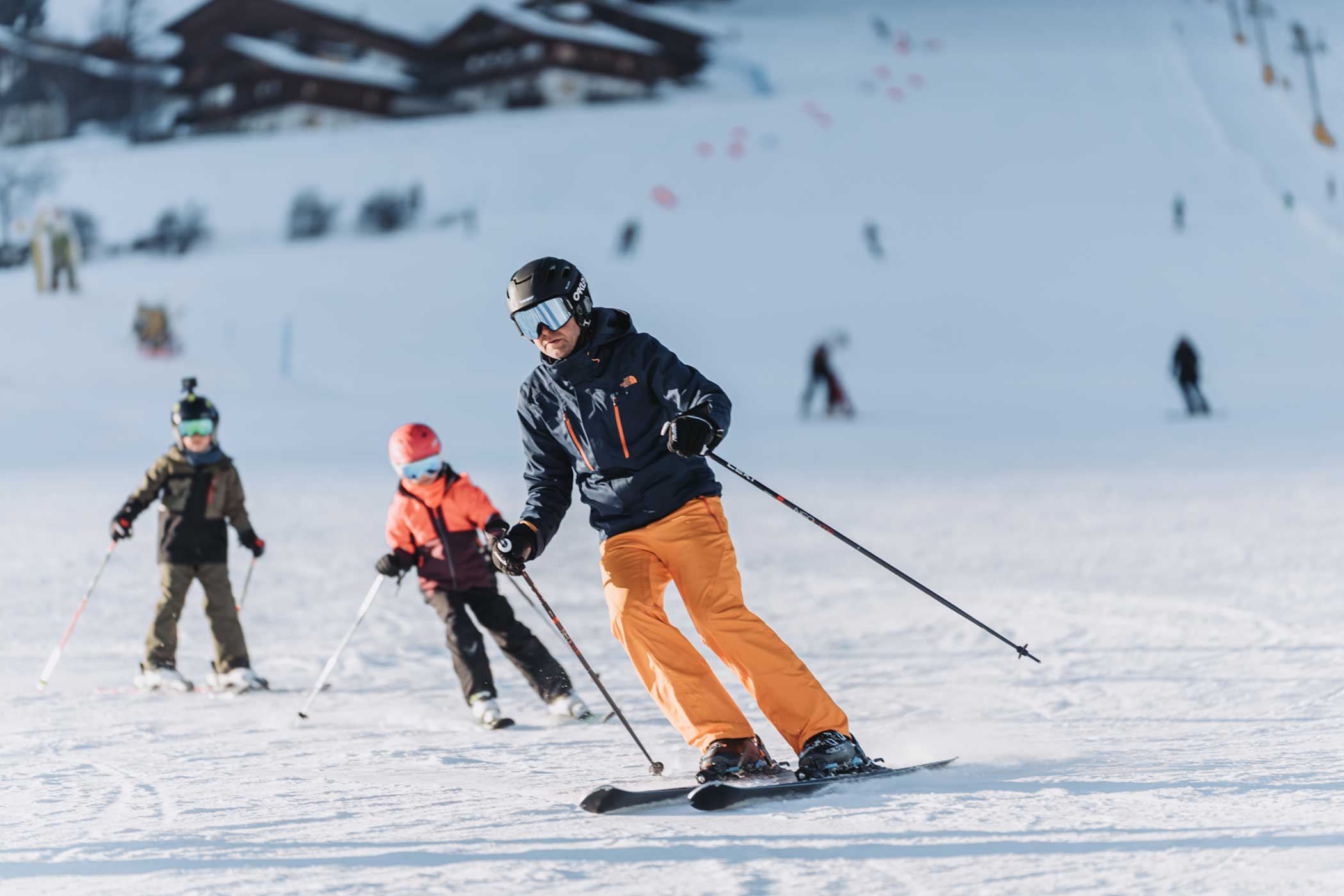 Drei Personen, die mit Helm und Winterausrüstung auf einer verschneiten Piste abfahren, mit Gebäuden im Hintergrund. - Hotel Waldheim