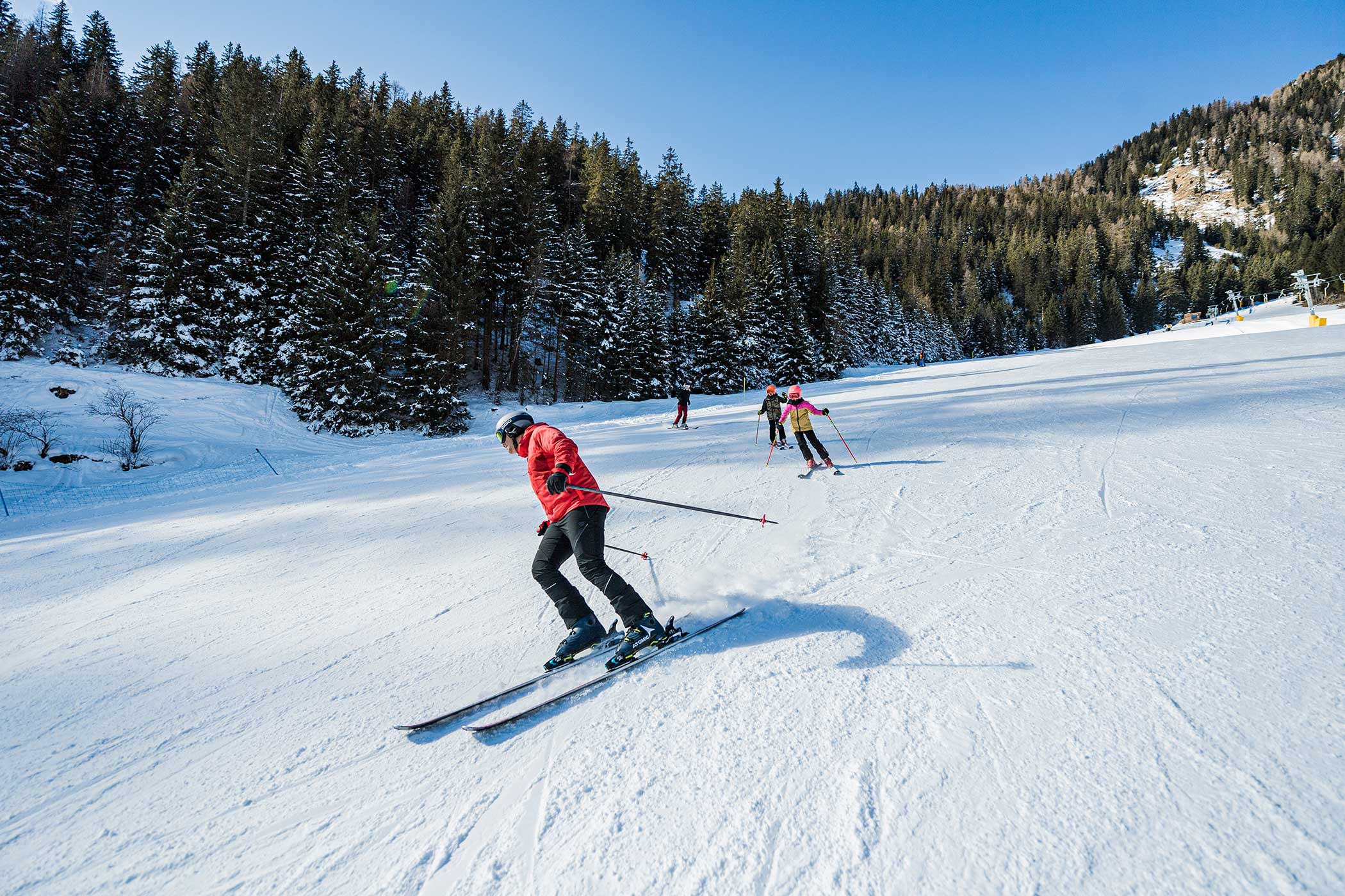 Skifahrer in farbenfrohen Jacken fahren auf einer verschneiten Piste, umgeben von Kiefern und blauem Himmel, bergab. - Hotel Waldheim