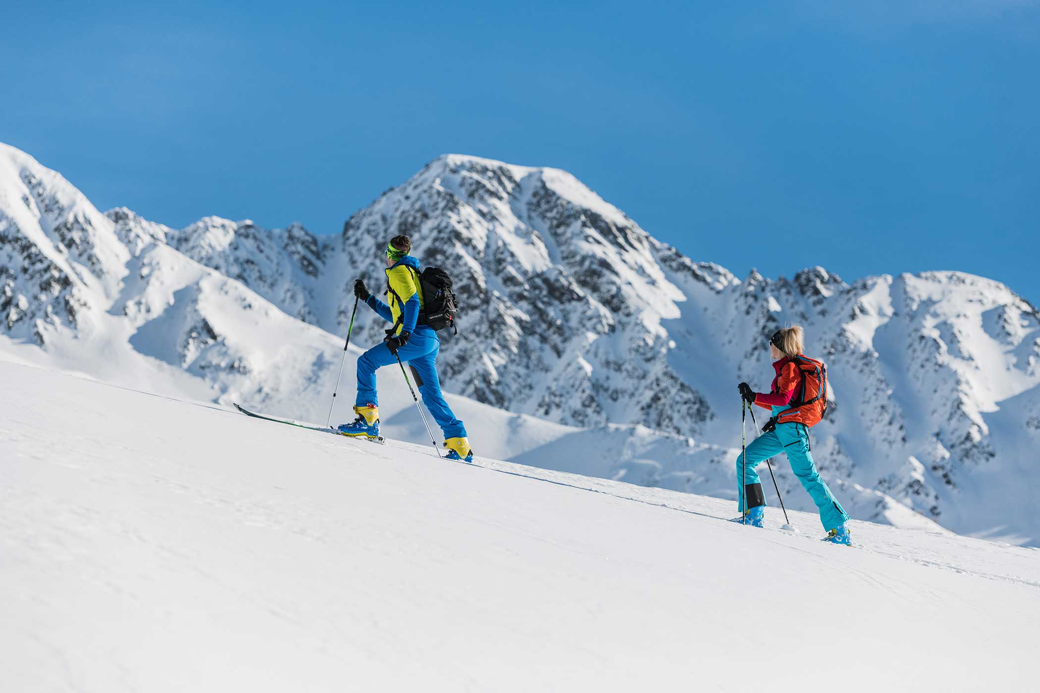 Zwei Personen in farbenfroher Kleidung fahren bergauf auf verschneiten Berghängen mit großen Gipfeln im Hintergrund. - Hotel Waldheim