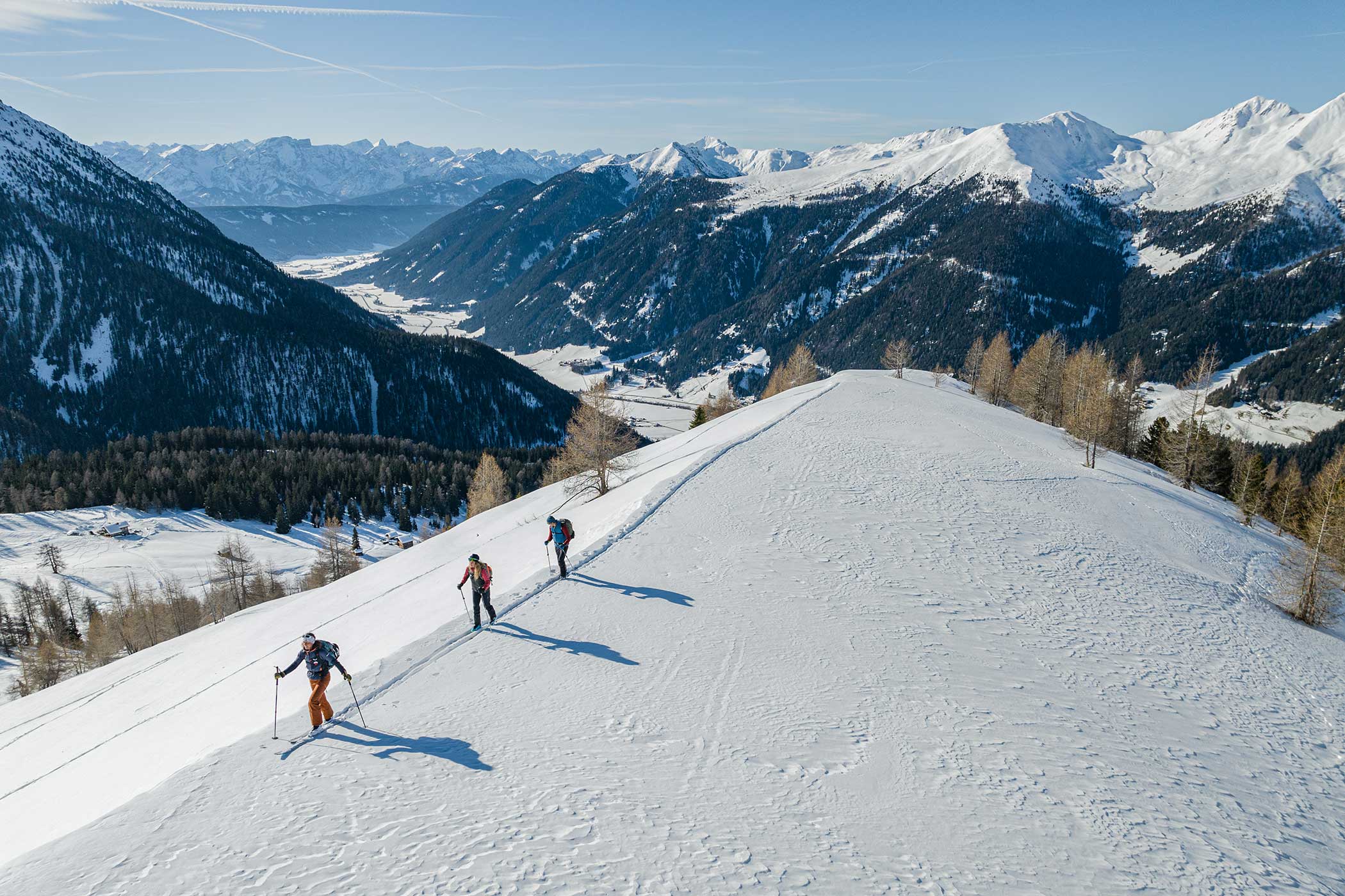 Drei Personen wandern mit Trekkingstöcken einen verschneiten Berghang hinauf, umgeben von schneebedeckten Gipfeln und Bäumen. - Hotel Waldheim