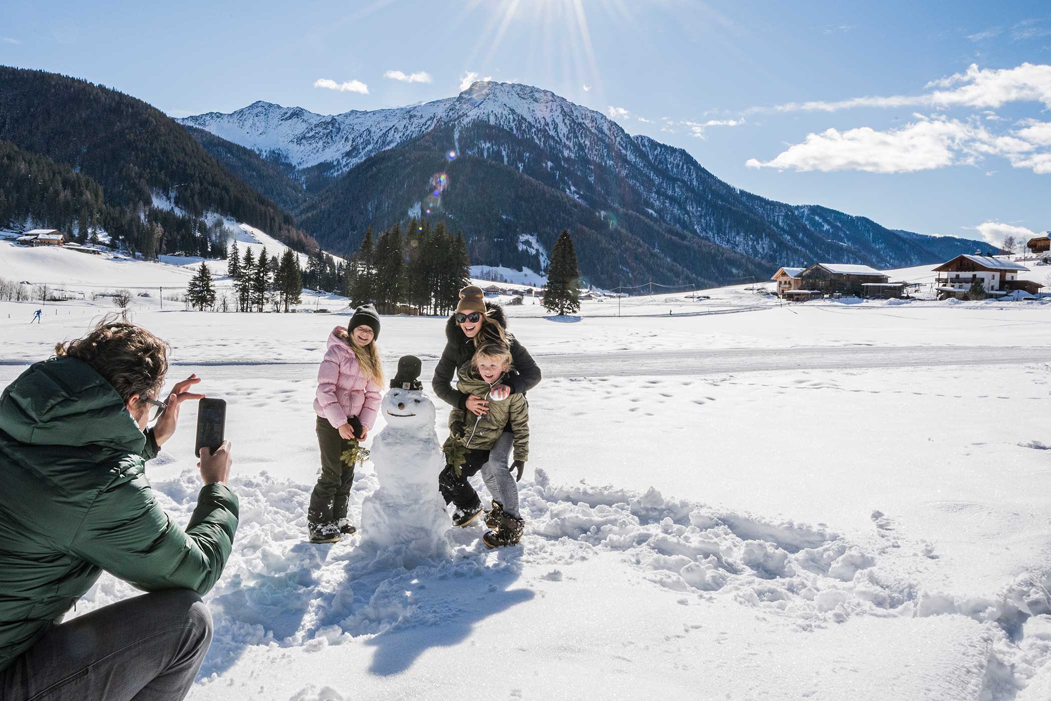 Eine Familie posiert neben einem Schneemann, während jemand ein Foto in einer verschneiten Berglandschaft unter einem sonnigen Himmel macht. - Hotel Waldheim