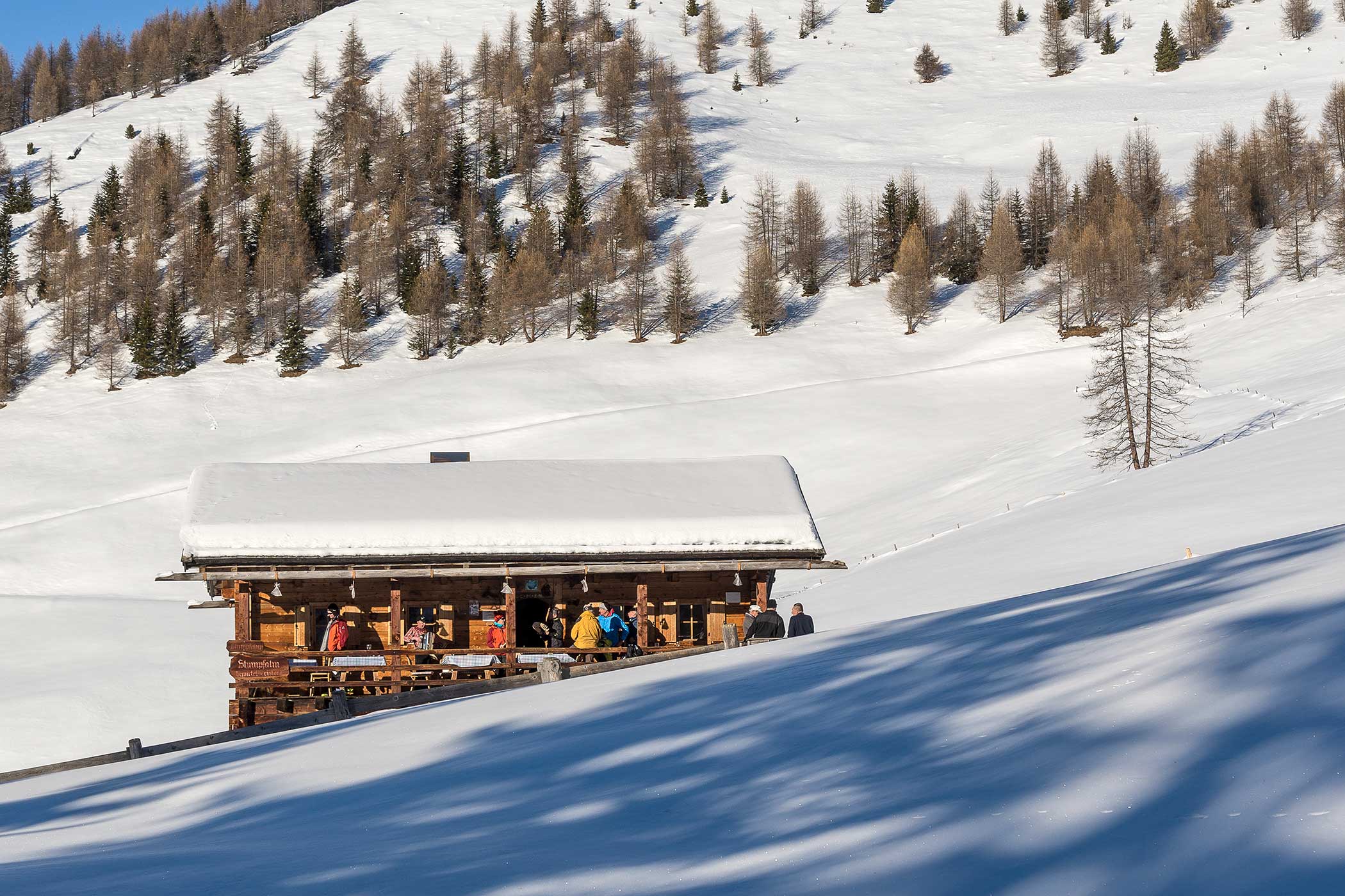 Eine Holzhütte mit Menschen draußen, umgeben von Schnee und verstreuten Bäumen an einem sonnigen Hang. - Hotel Waldheim