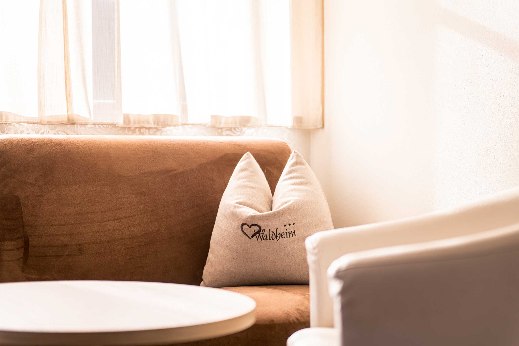 Sonnenbeschienenes, gemütliches Sofa mit einem weißen, mit Waldbrein bestickten Kissen, daneben ein weißer Stuhl und ein runder Tisch. - Hotel Waldheim
