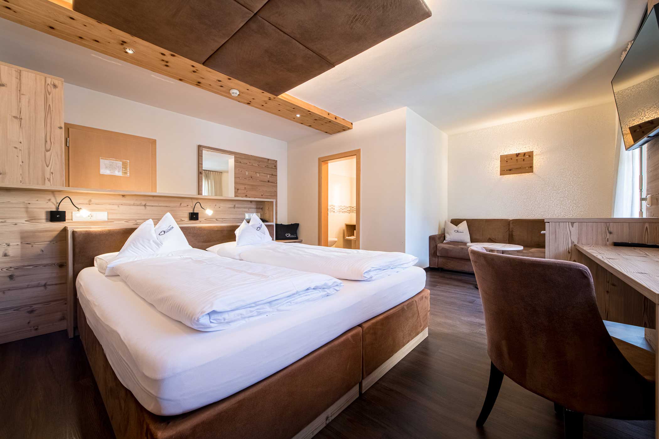 Modernes Hotelzimmer mit zwei Betten, Holzmöbeln, einem Schreibtisch, einem Sofa und warmer Beleuchtung. - Hotel Waldheim