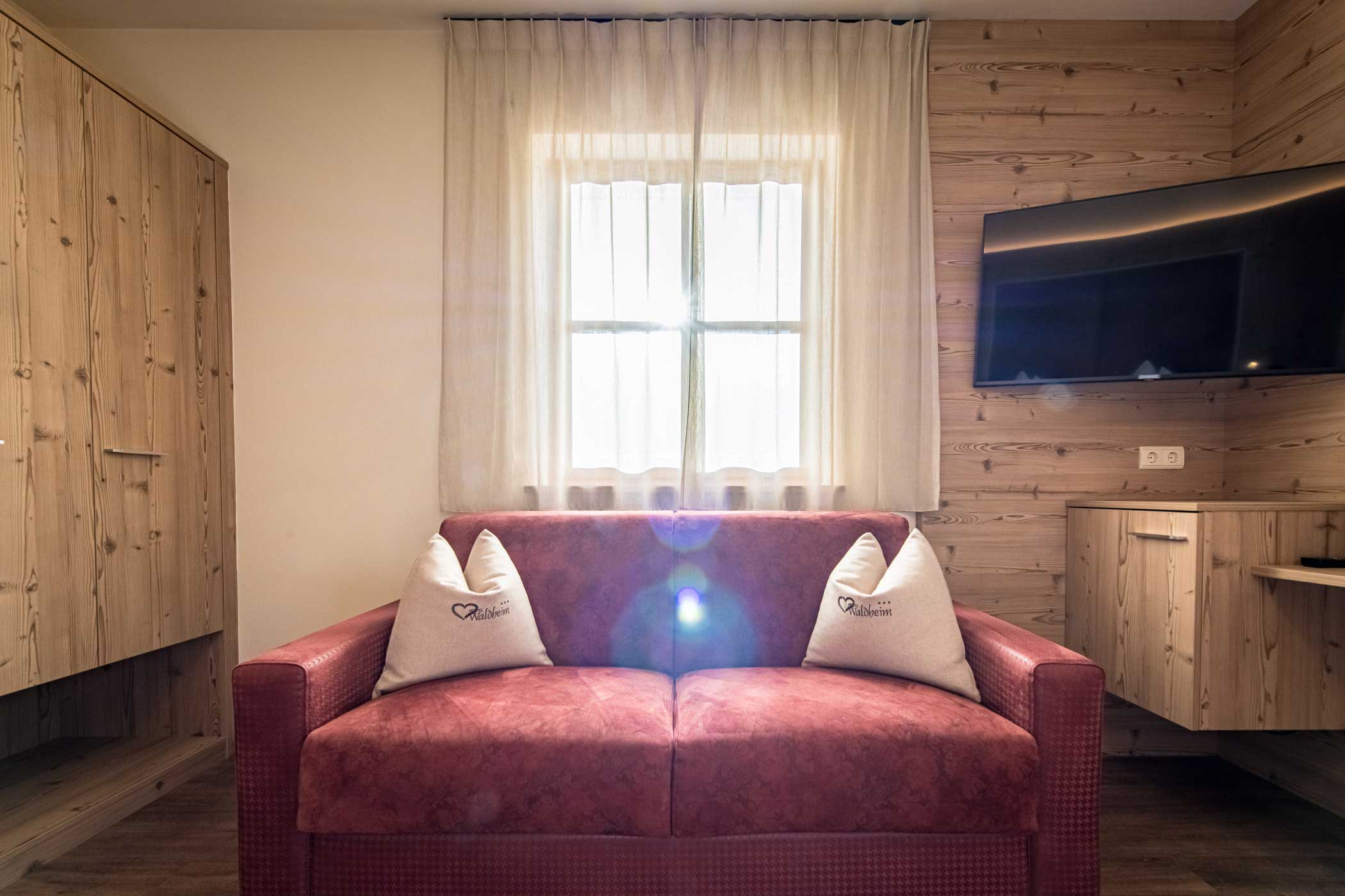 Ein rotes Sofa mit zwei Kissen steht vor einem Fenster, an der holzgetäfelten Wand ist ein Fernseher angebracht. - Hotel Waldheim