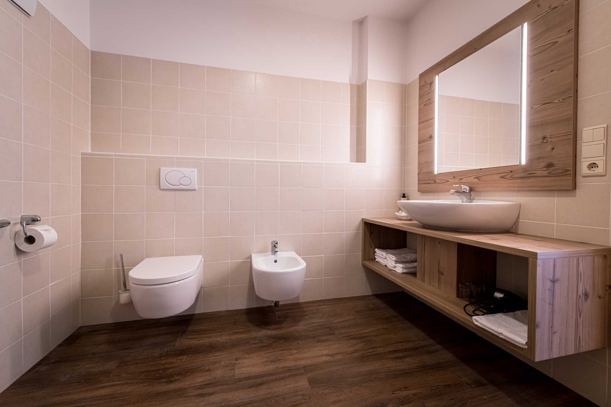 Modernes Badezimmer mit wandmontierter Toilette und Bidet, Holzwaschtisch, Gefäßwaschbecken und großem Spiegel. - Hotel Waldheim