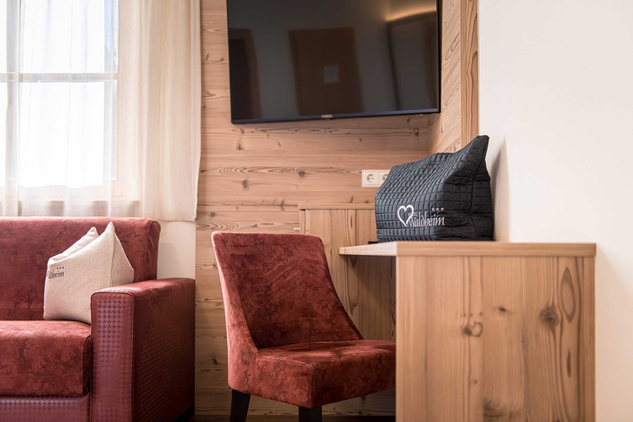 Ein roter Sessel und ein rotes Sofa neben einem hölzernen Schreibtisch und einem Fernseher, durch ein vorgehängtes Fenster fällt Sonnenlicht. - Hotel Waldheim