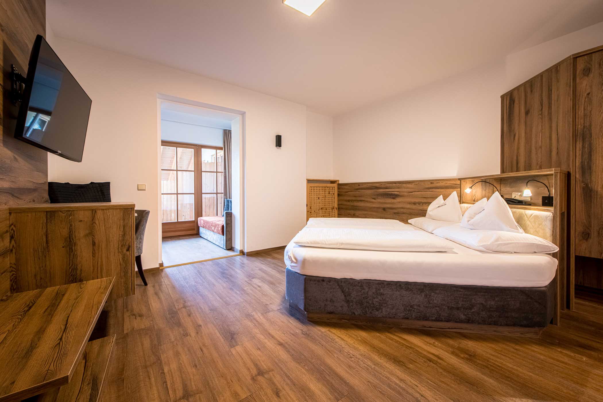 Modernes Hotelzimmer mit Doppelbett, Holzfußboden, TV, Schreibtisch und einer sonnenbeschienenen Sitzecke an großen Fenstern. - Hotel Waldheim